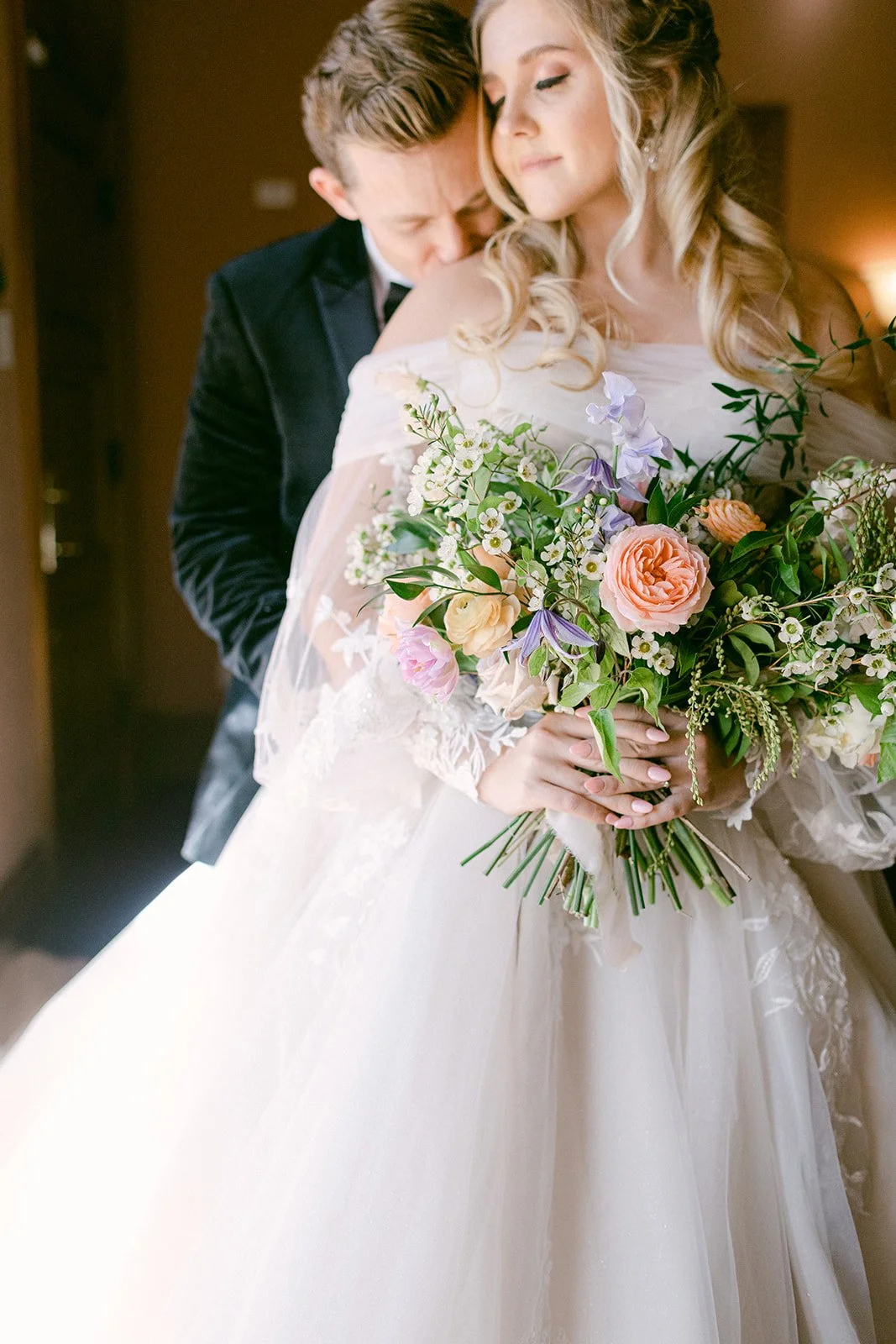 A bride in a white wedding dress holding a large bouquet of flowers, with a groom standing behind her, both with smiles and eyes closed, showing intimacy and love.