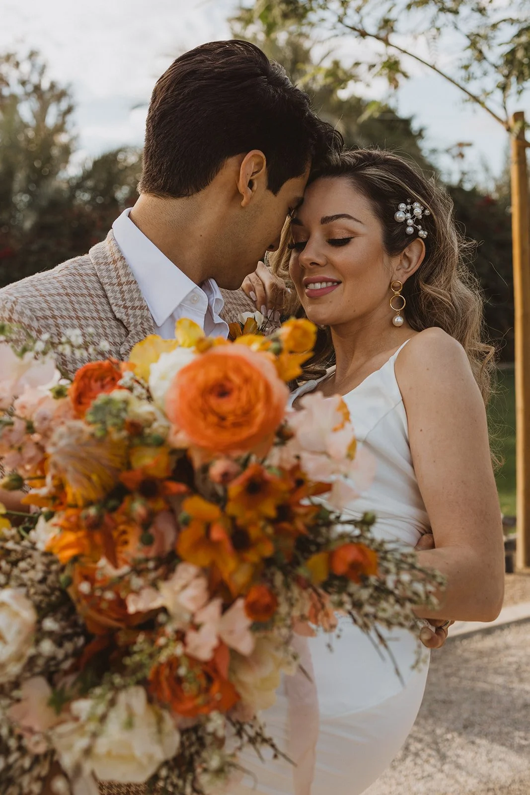 A bride and groom share a tender moment outdoors during their wedding, with the bride holding a large bouquet of colorful flowers and both with their foreheads touching.