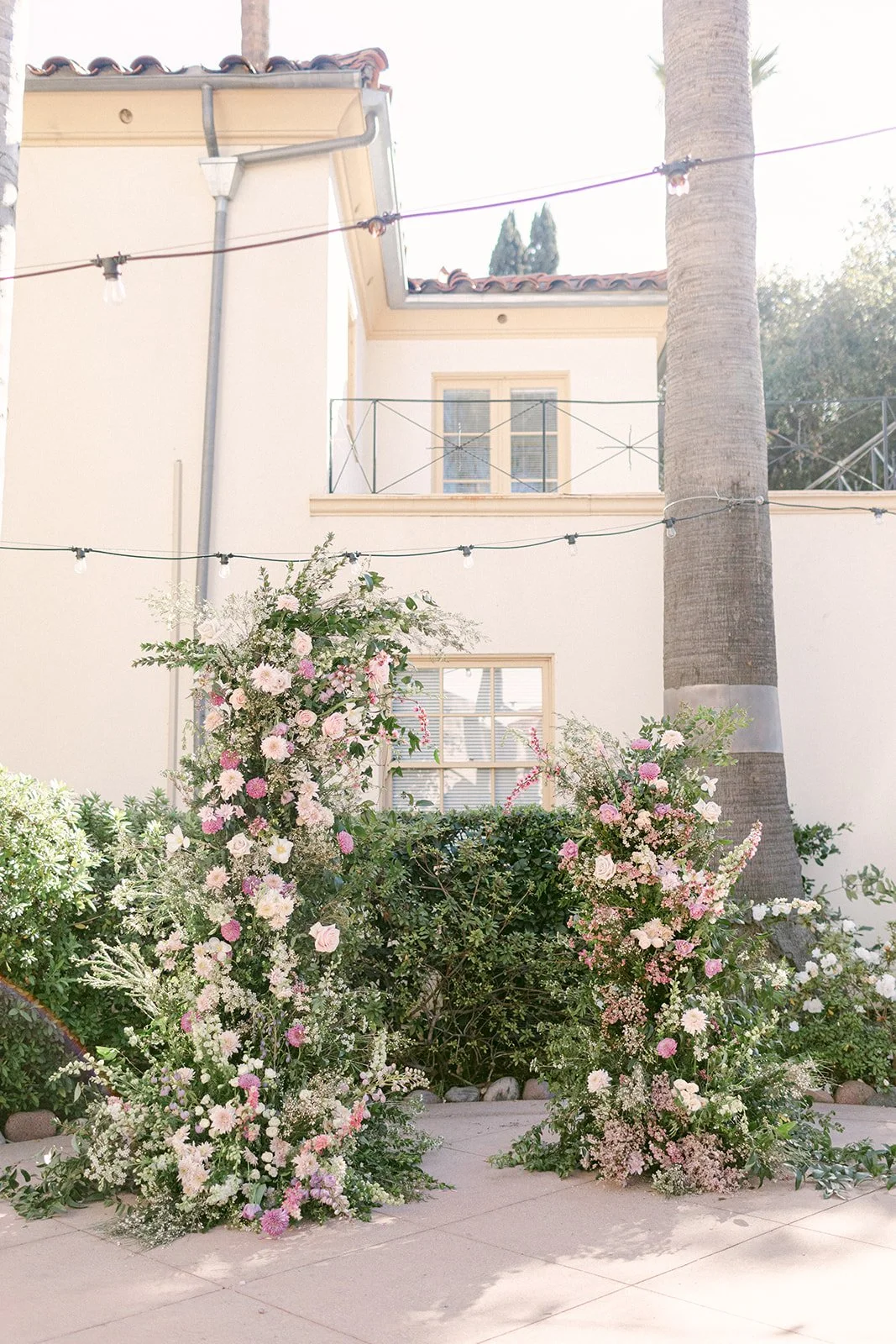 Decorative floral arrangements with pink and white flowers outdoors near a house with string lights hanging overhead.