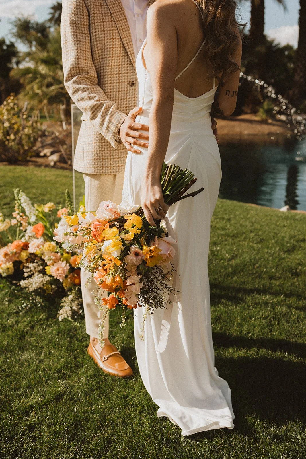 A bride is holding a colorful bouquet of flowers, standing outdoors near a body of water, with a person in a checkered blazer visible behind her.