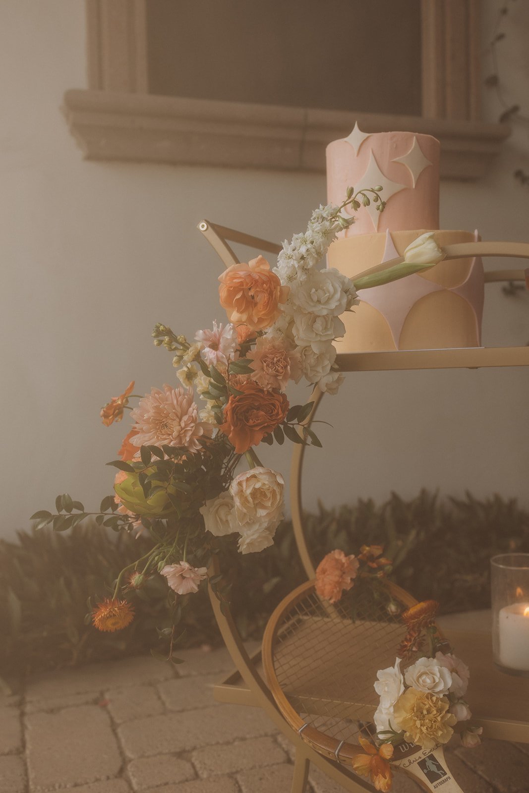 A pink, layered, decorative cake with star-shaped accents on a gold metal stand, adorned with fresh flowers including roses, carnations, and peonies, on a table with a lit candle in the background.