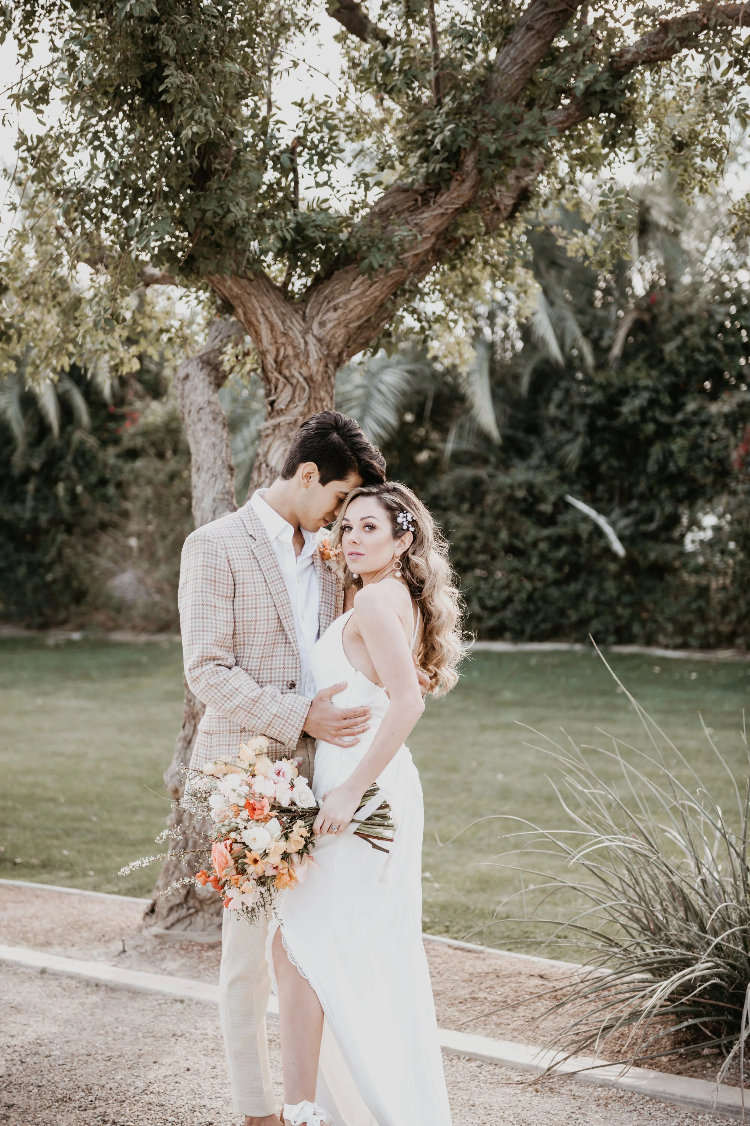 A bride and groom standing outdoors near a large tree, with the groom embracing the bride as they look into each other's eyes. The bride holds a bouquet of flowers. The background includes lush greenery and a clear sky.