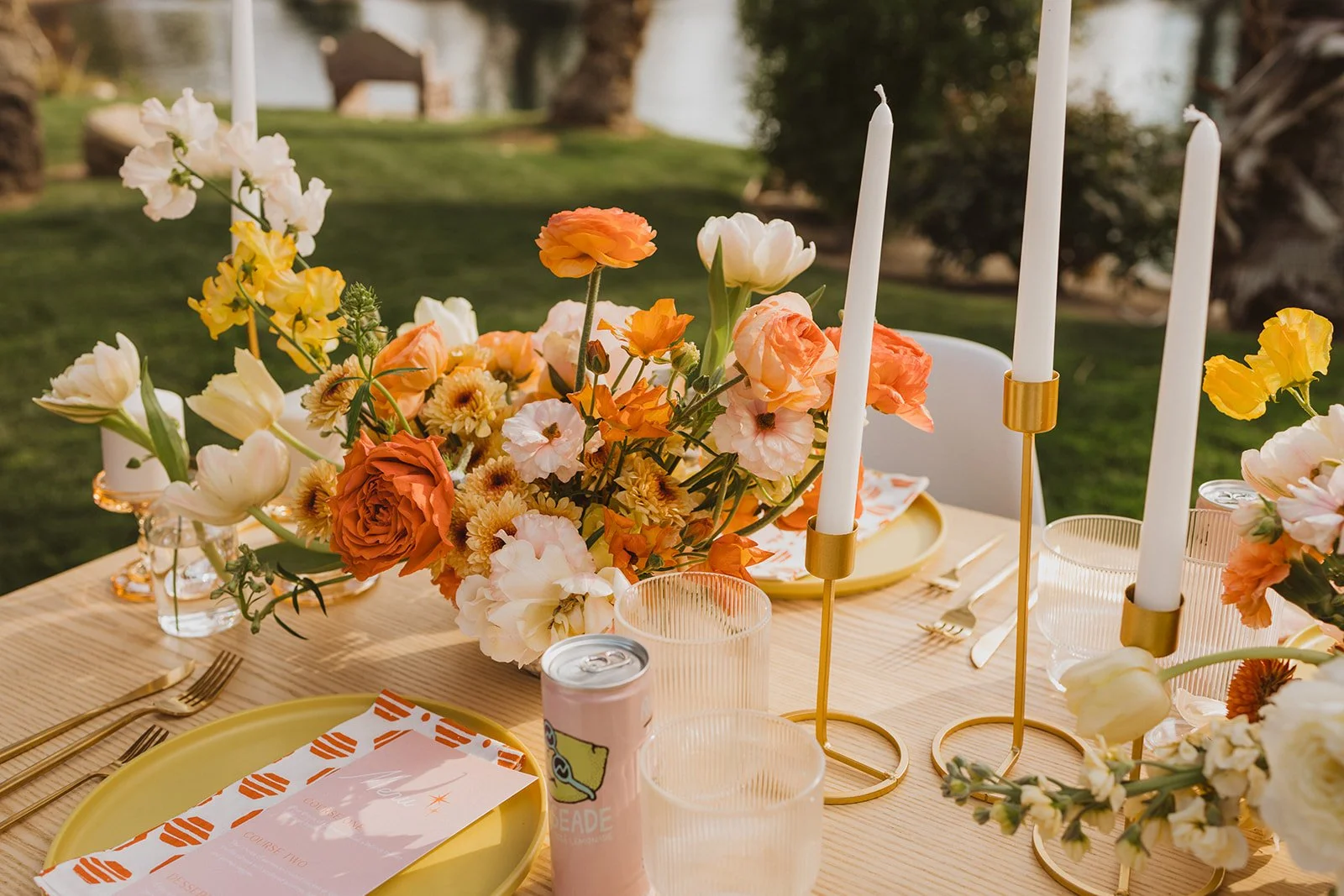 A table decorated with a floral centerpiece, white candles in gold holders, a pink drink, a soda, yellow plates, and orange and white napkins, set outdoors on green grass near water.