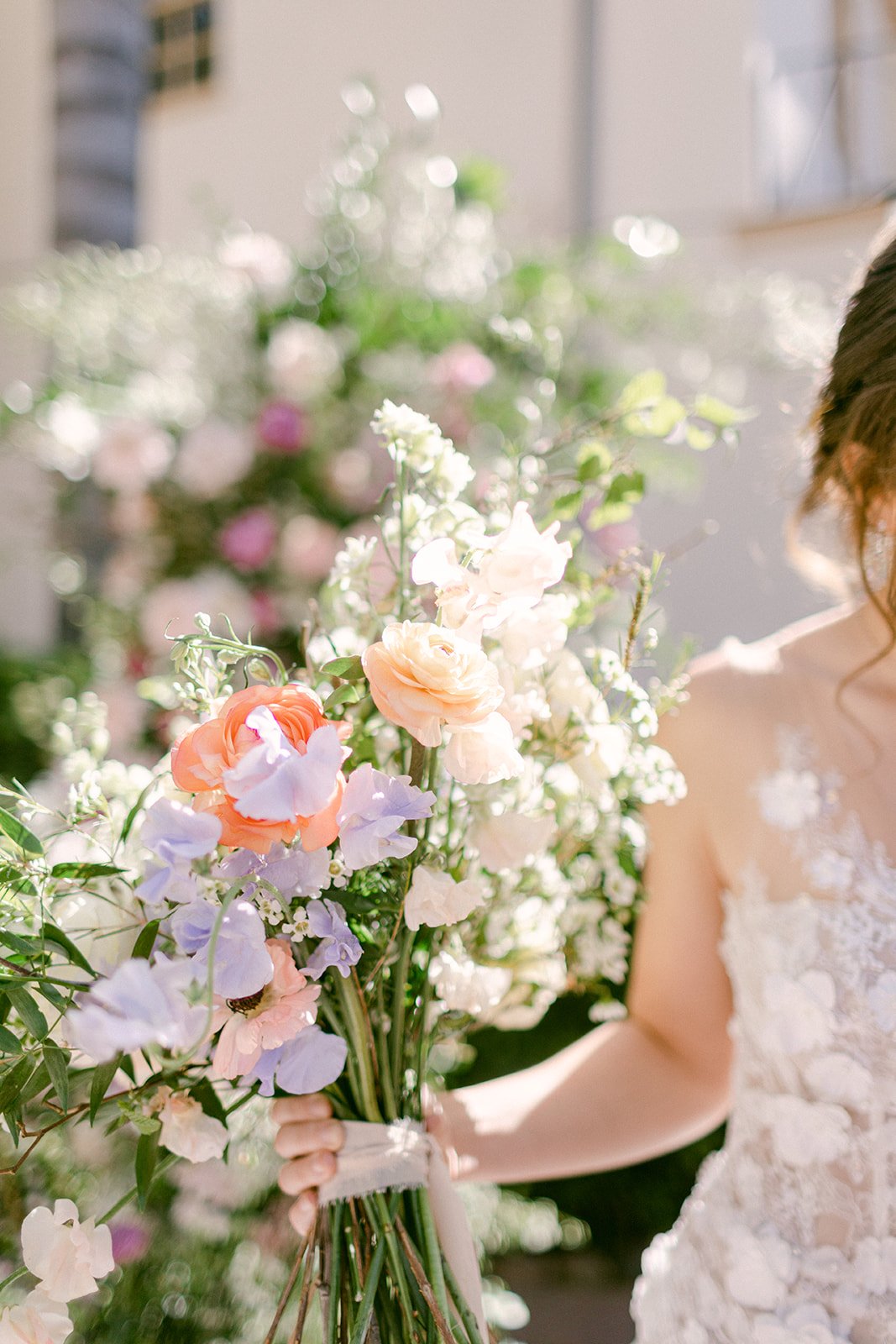 A person in a wedding dress holds a bouquet of pastel-colored flowers, with a blurred background of more flowers.