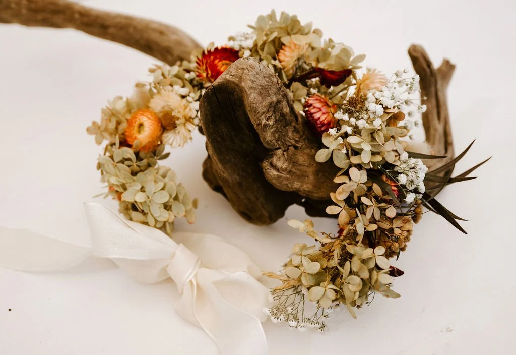 A floral crown made of dried flowers and light-colored greenery, with a white ribbon tied in a bow.