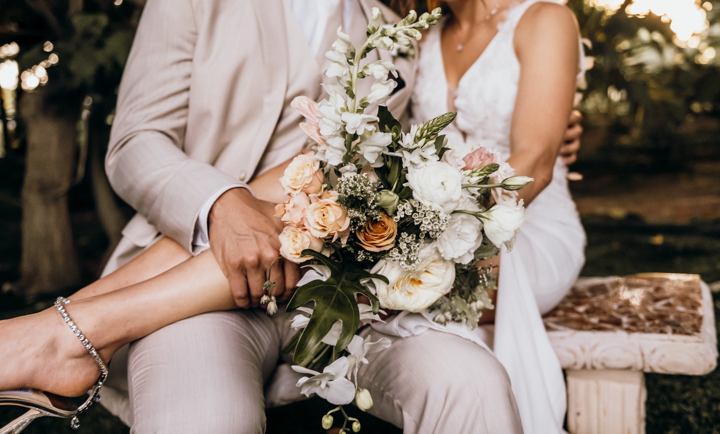 A couple holding a bouquet of white, cream, and peach flowers during a wedding, sitting outdoors with trees and sunlight in the background.