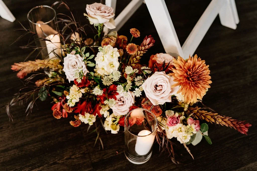 A floral arrangement with roses, chrysanthemums, and various autumn-colored flowers, accompanied by a lit white candle in a glass holder, set on a dark wooden surface.