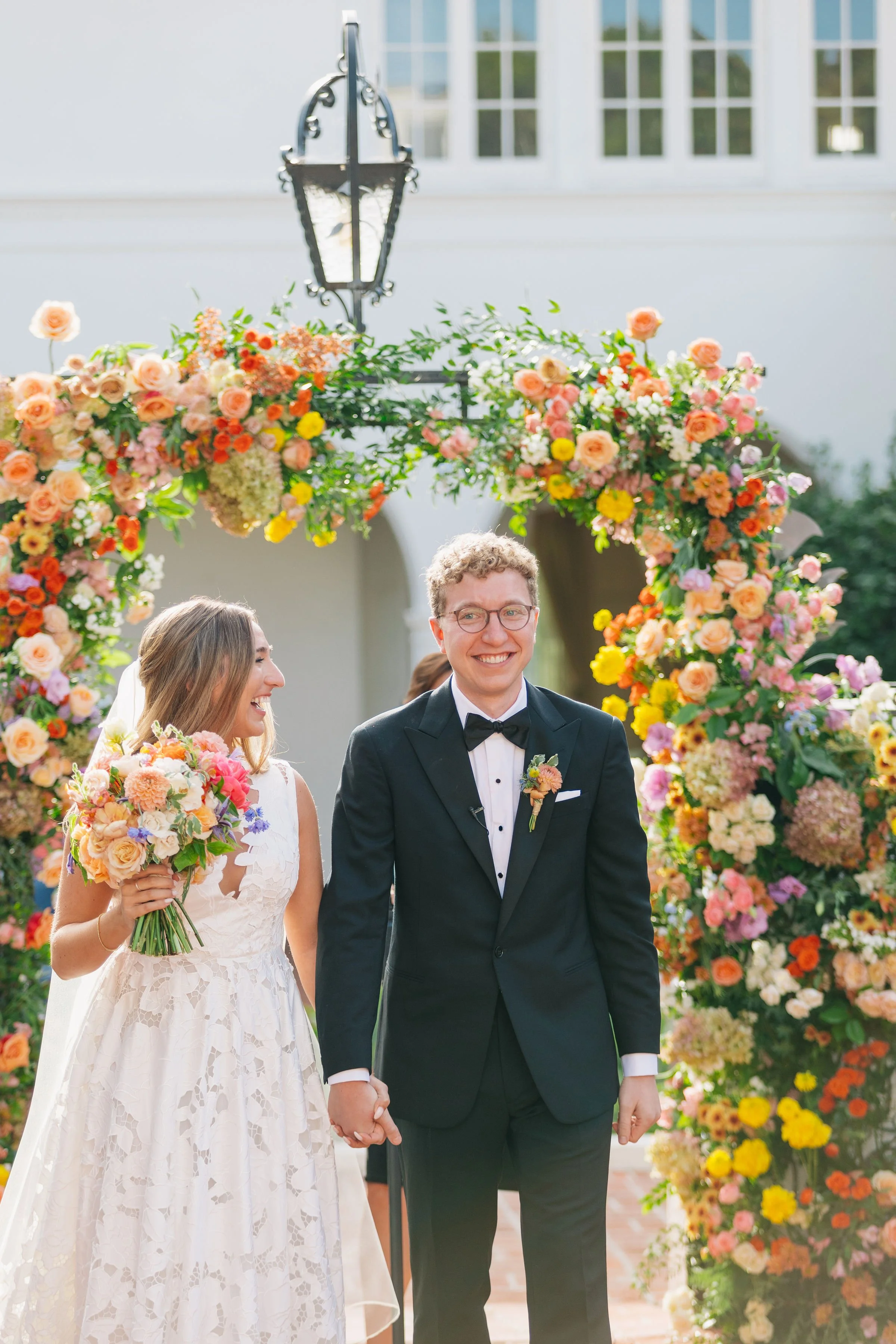 A wedding couple holding hands in front of a floral arch, with a house and window in the background, smiling during their outdoor wedding ceremony.