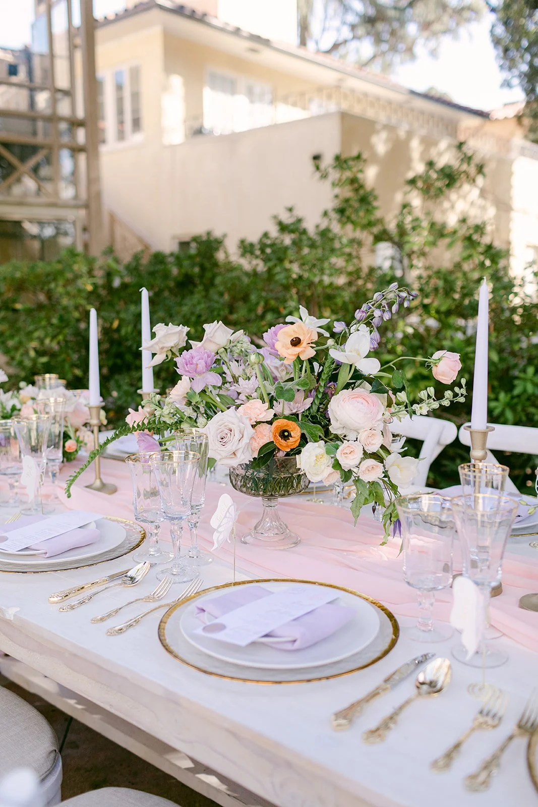 A decorated outdoor table with a pink table runner, fine china, gold utensils, tall pink candles, and a floral centerpiece with white, purple, and peach flowers.