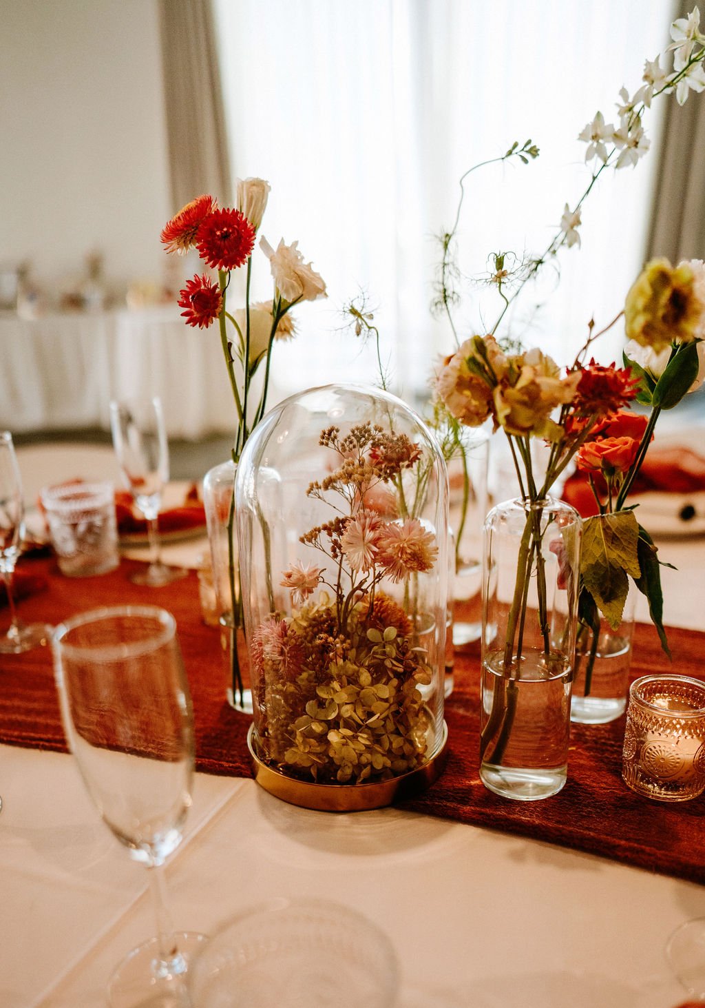 Elegant table centerpiece with various flowers in glass vases and a glass dome filled with dried flowers, on a table with a red table runner, surrounded by glasses and candles, in a well-lit room.