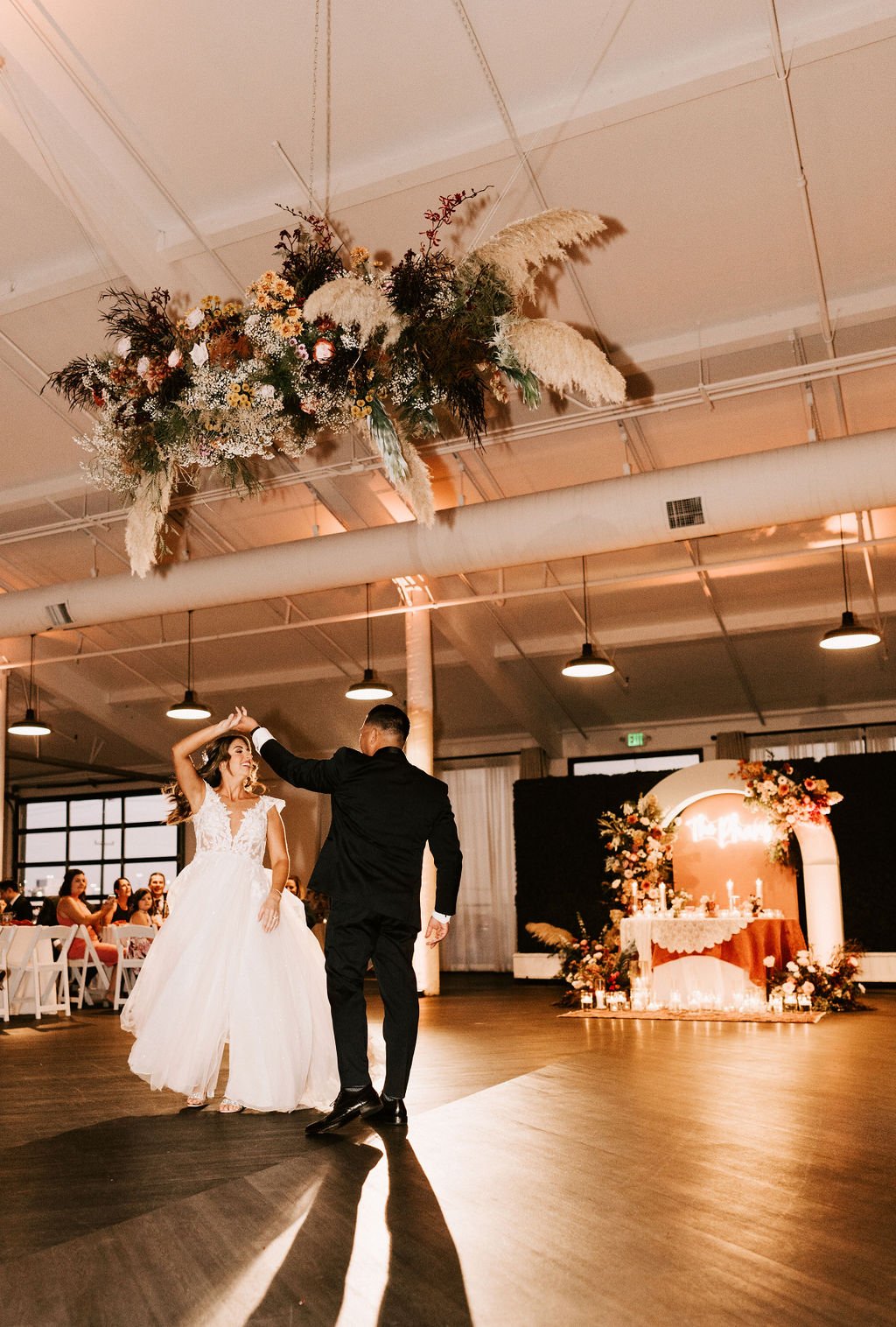 A bride and groom dancing at their wedding reception with a floral installation overhead and a decorated altar in the background.