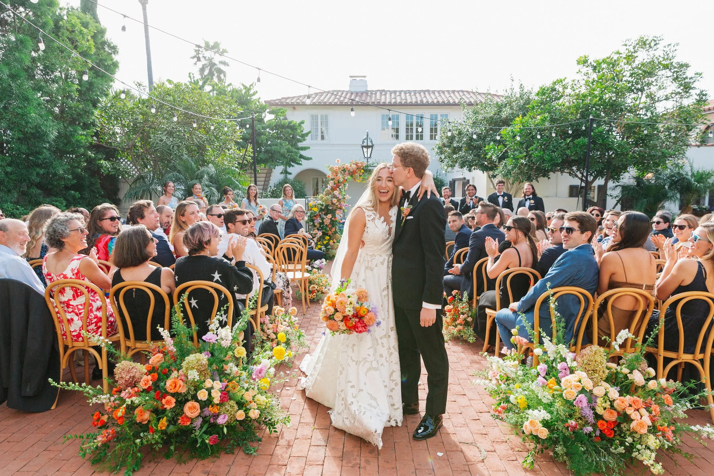 A wedding ceremony outdoors with the bride and groom embracing and kissing on a brick walkway, surrounded by floral arrangements and seated guests, under string lights and a large arch of colorful flowers, with a white building and trees in the backg