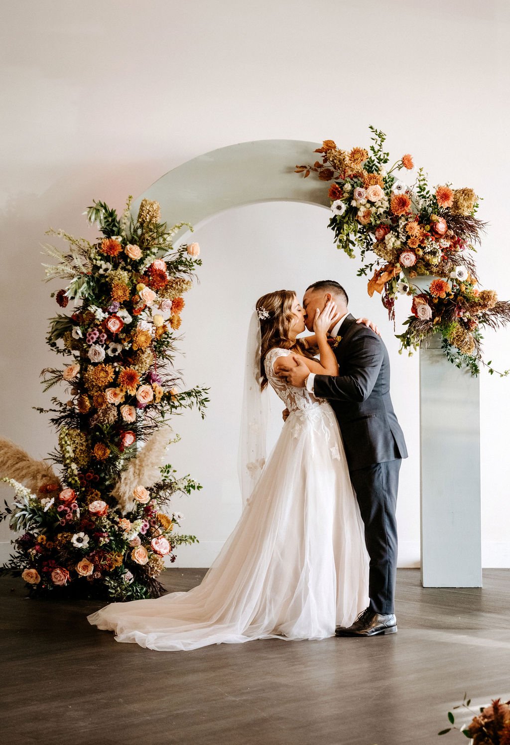 A bride and groom kiss during their wedding ceremony, standing under a floral arch adorned with pink, peach, and white flowers and greenery.