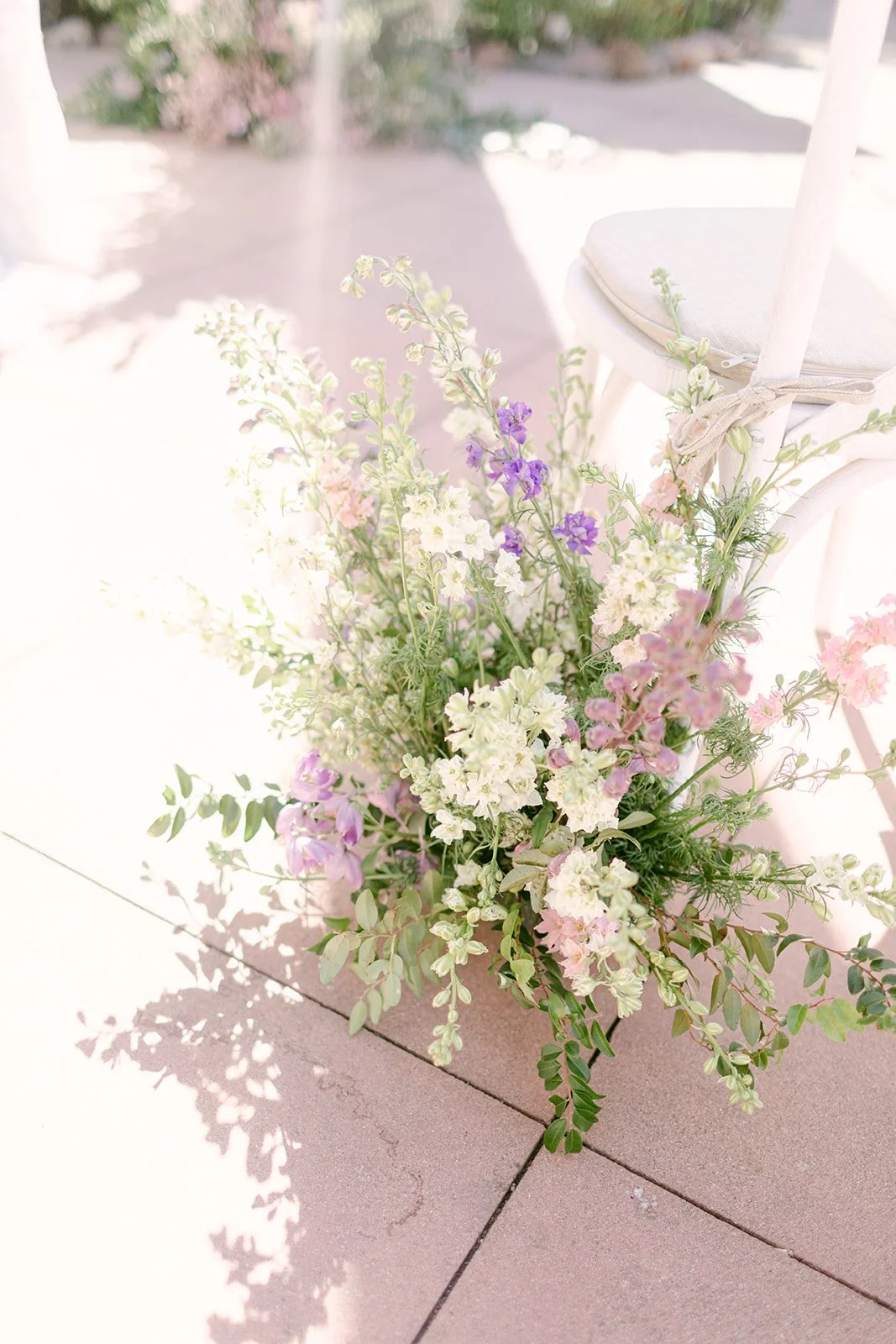A bouquet of mixed pink, white, purple and green flowers placed on a pinkish outdoor tiled surface near a white chair with a fabric covering and tied with a bow.