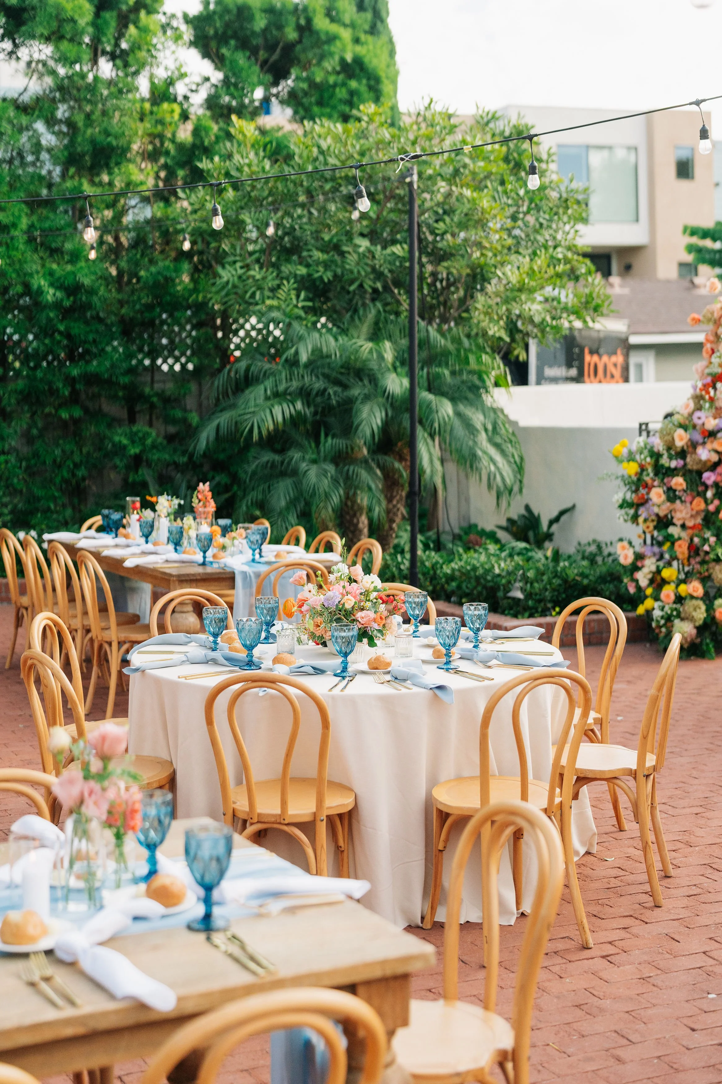 Outdoor dining area with round tables covered in white tablecloths, set with blue glassware, silverware, white napkins, and floral centerpieces. Wooden chairs surround the tables. String lights are hung overhead. The area is decorated with lush green