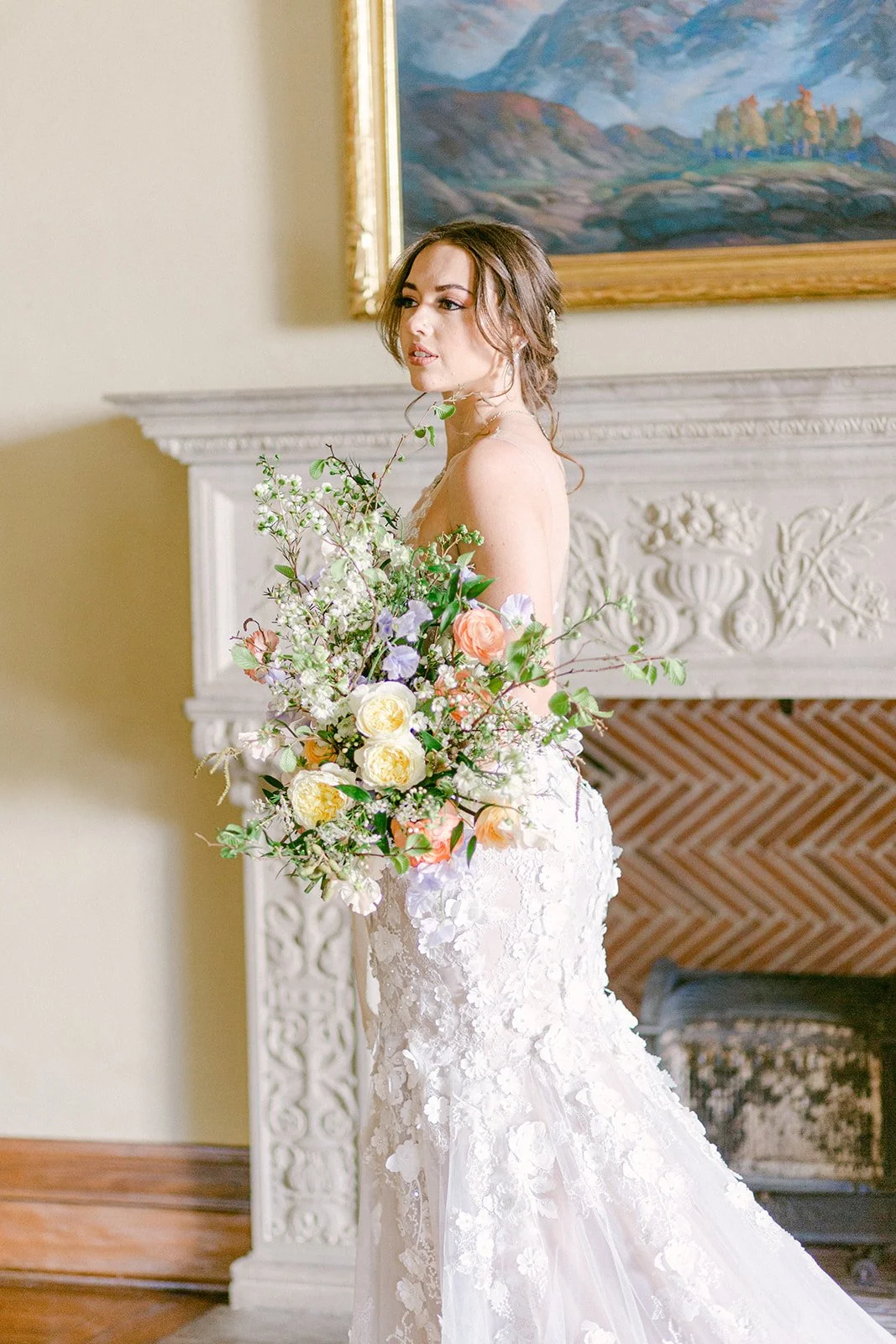 A bride in a white lace wedding dress holding a large bouquet of colorful flowers, standing indoors with an ornate fireplace and a landscape painting in the background.
