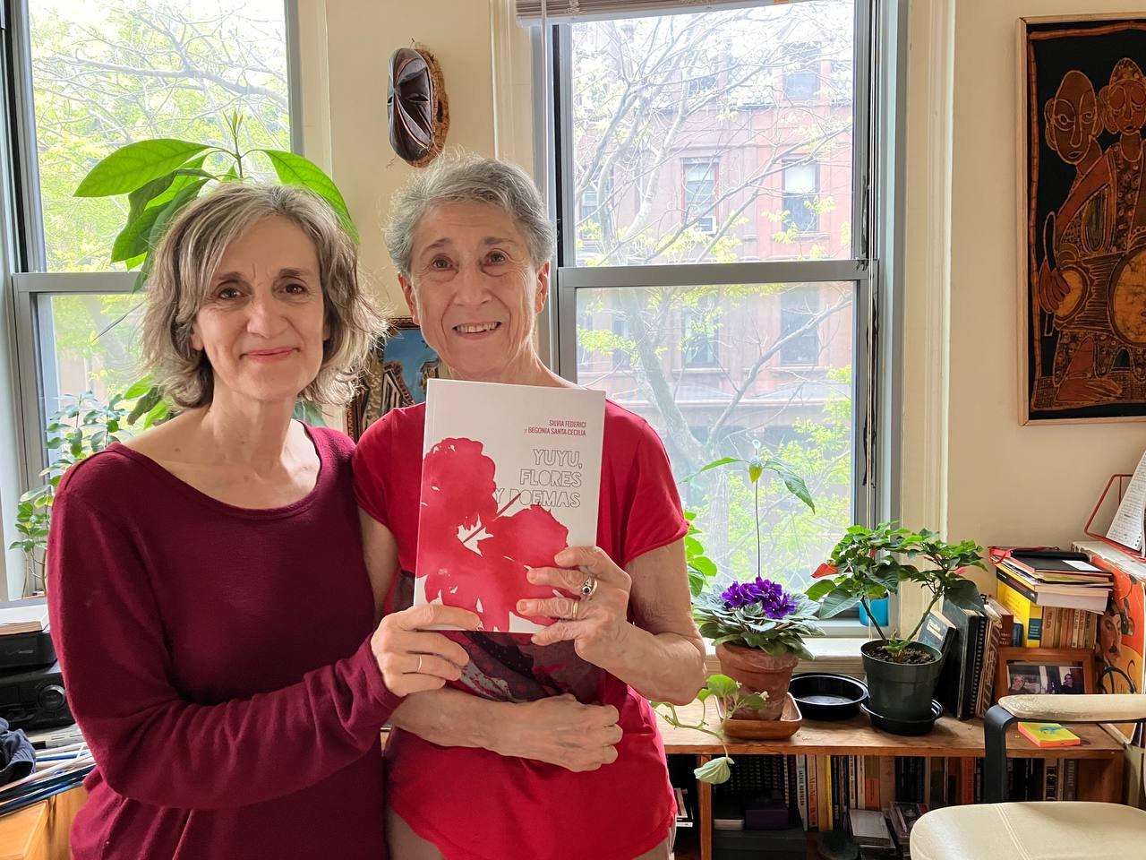 Begonia Santa-Cecilia y Silvia Federici muestran su libro 'Yuyu, flores y poemas'. Posan cercanas, en un ligero abrazo, ambas vestidas de tonos de rojo, que combinan con la portada. Están en un departamento iluminado por el sol, decorado con plantas.