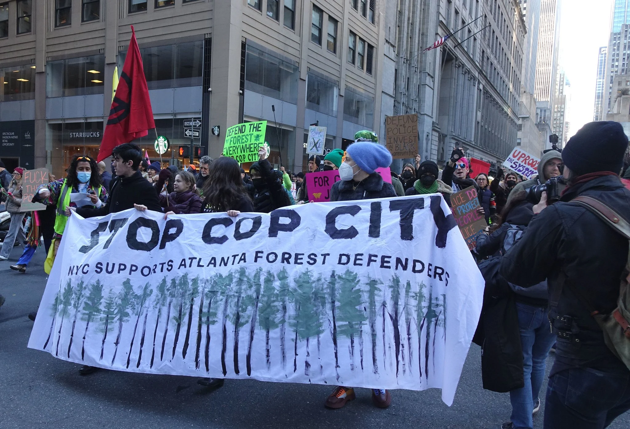 En una calle de Nueva York, tres jóvenes sostienen una manta que dice "Stop Cop City: la ciudad de Nueva York apoya a los defensores del bosque de Atlanta". Detrás , una multitud sostiene carteles con mensajes como "Desenraiza el sistema".