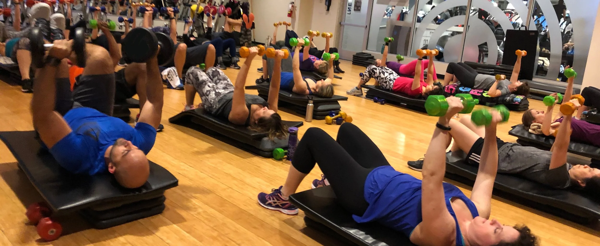 Group of people lying on their backs doing a bench press with dumbbells in a group fitness class