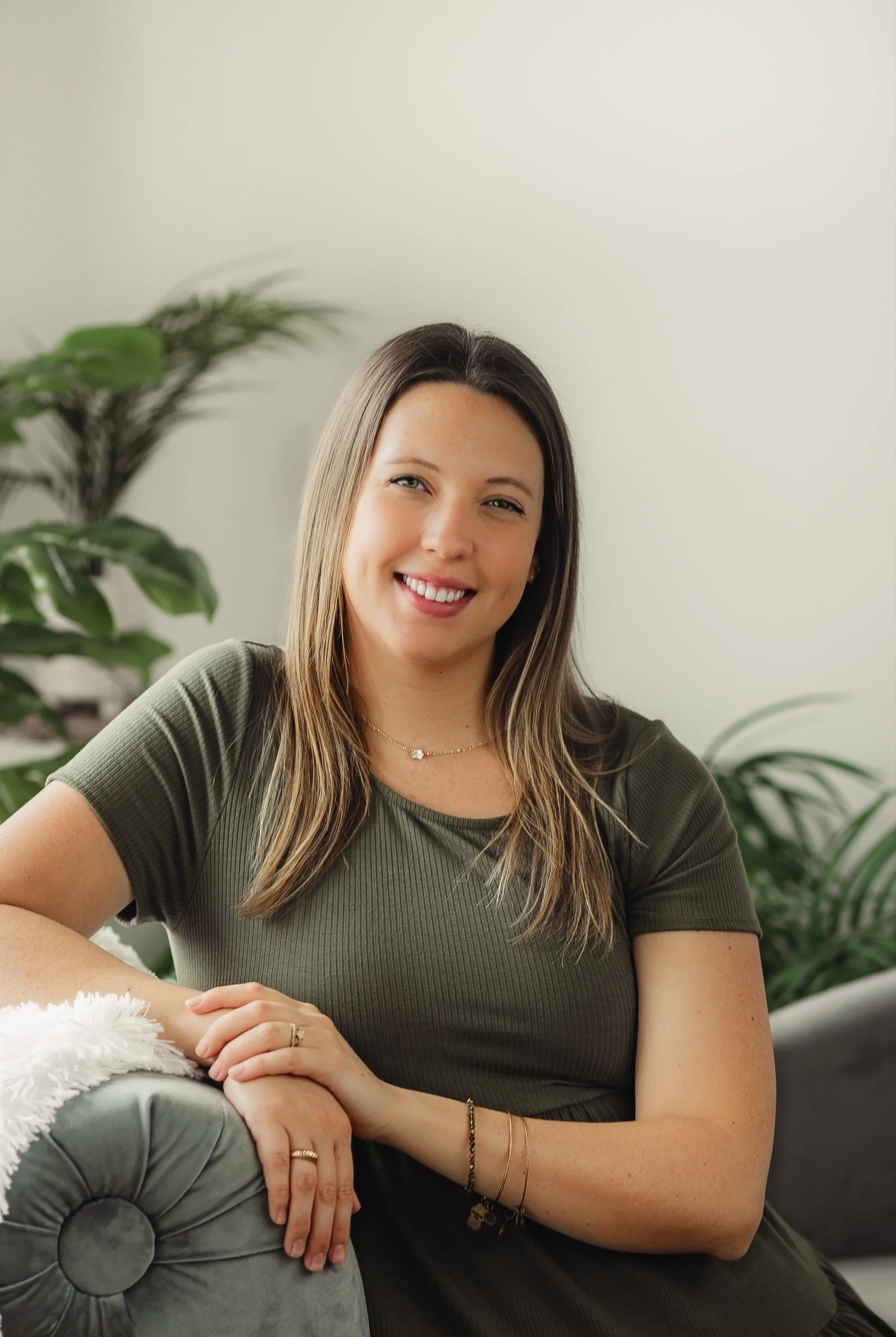 A smiling woman sitting indoors on a gray sofa, wearing an olive green shirt, with plants in the background.