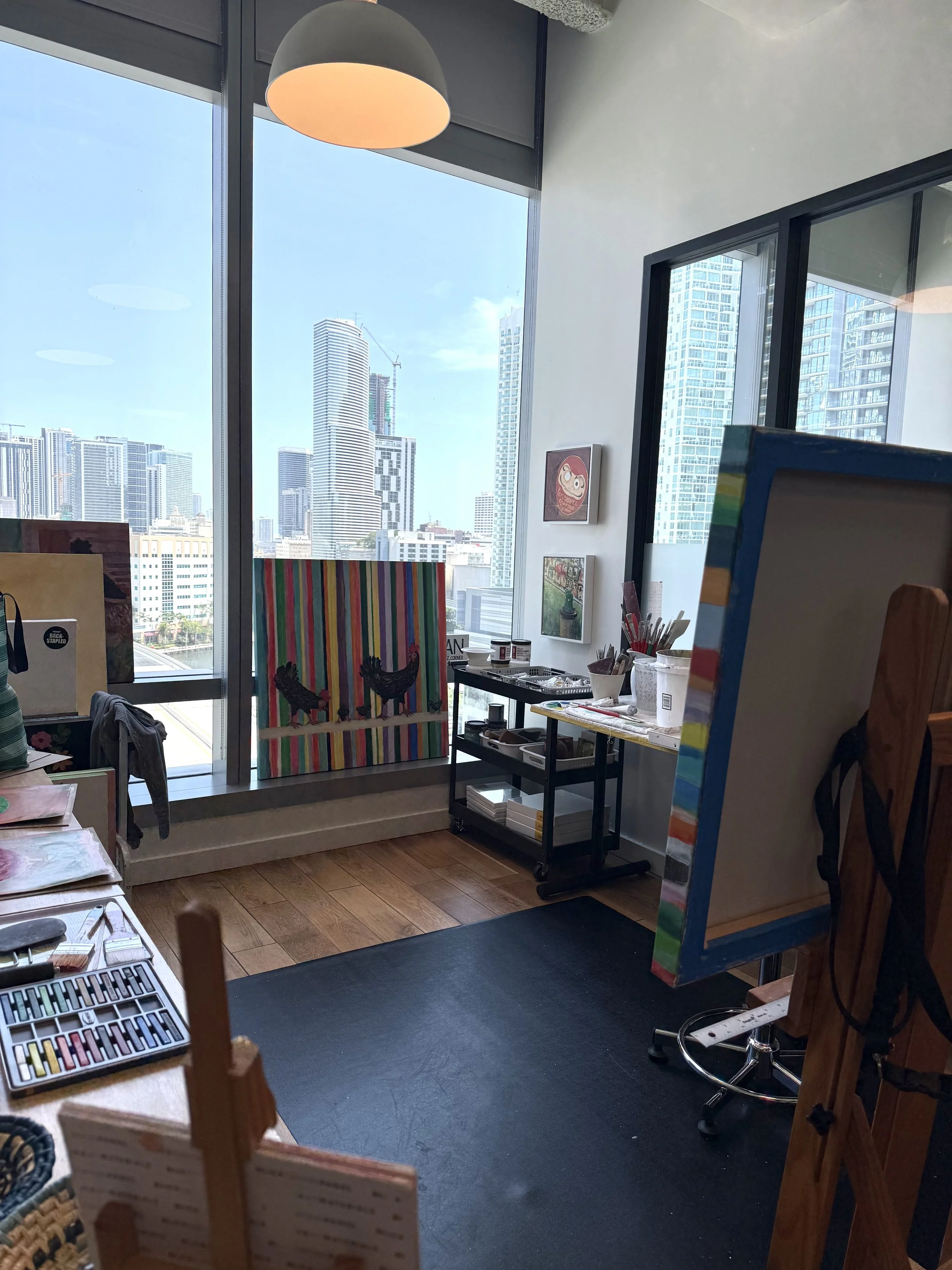 Interior of an art studio with large windows showing a city skyline with tall buildings and a crane. The room contains art supplies, a colorful canvas, a stool, and paintings on the wall.