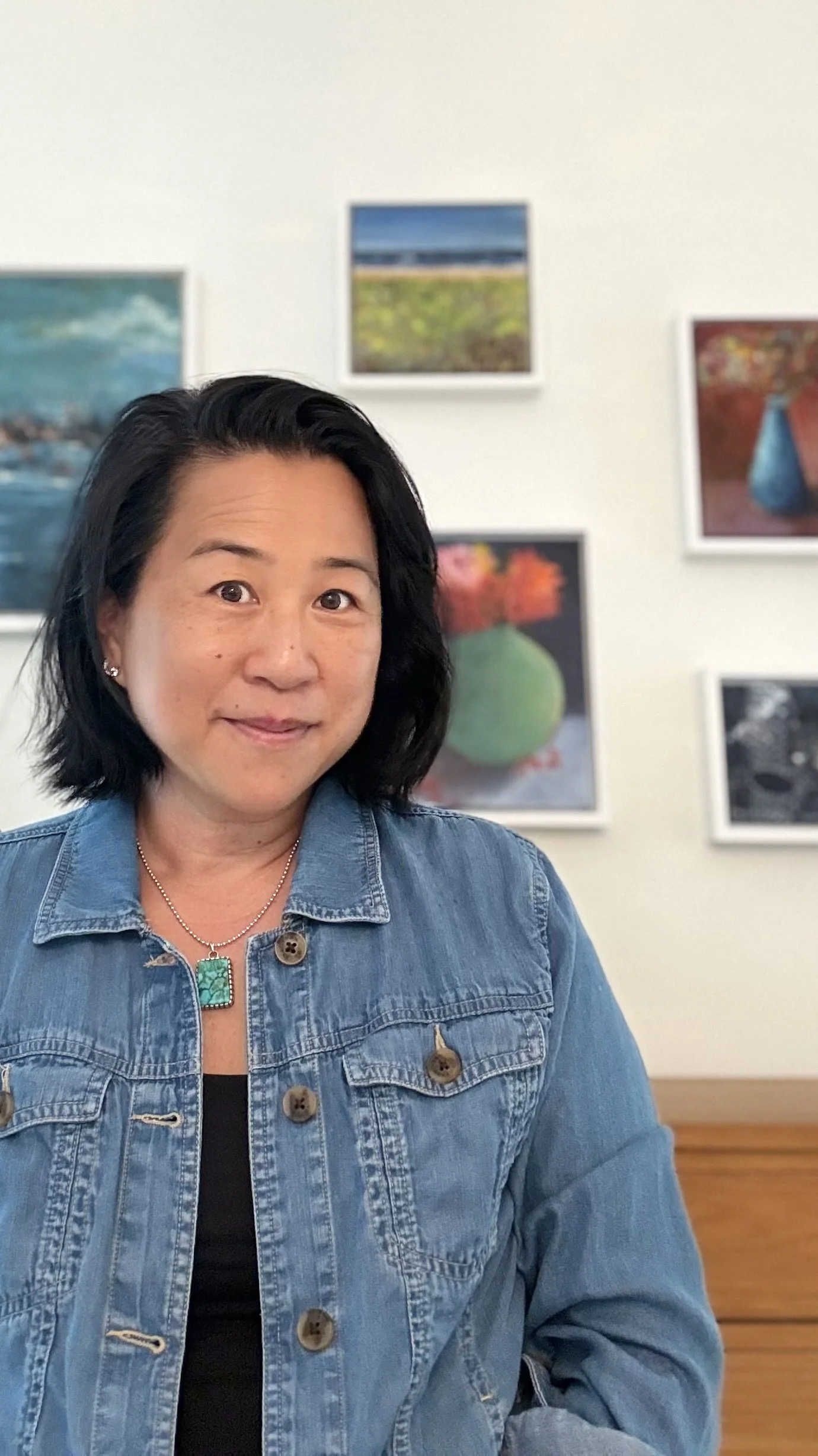 A woman with short black hair wearing a denim jacket with a turquoise pendant necklace. She is standing in front of a wall with framed landscape photos.