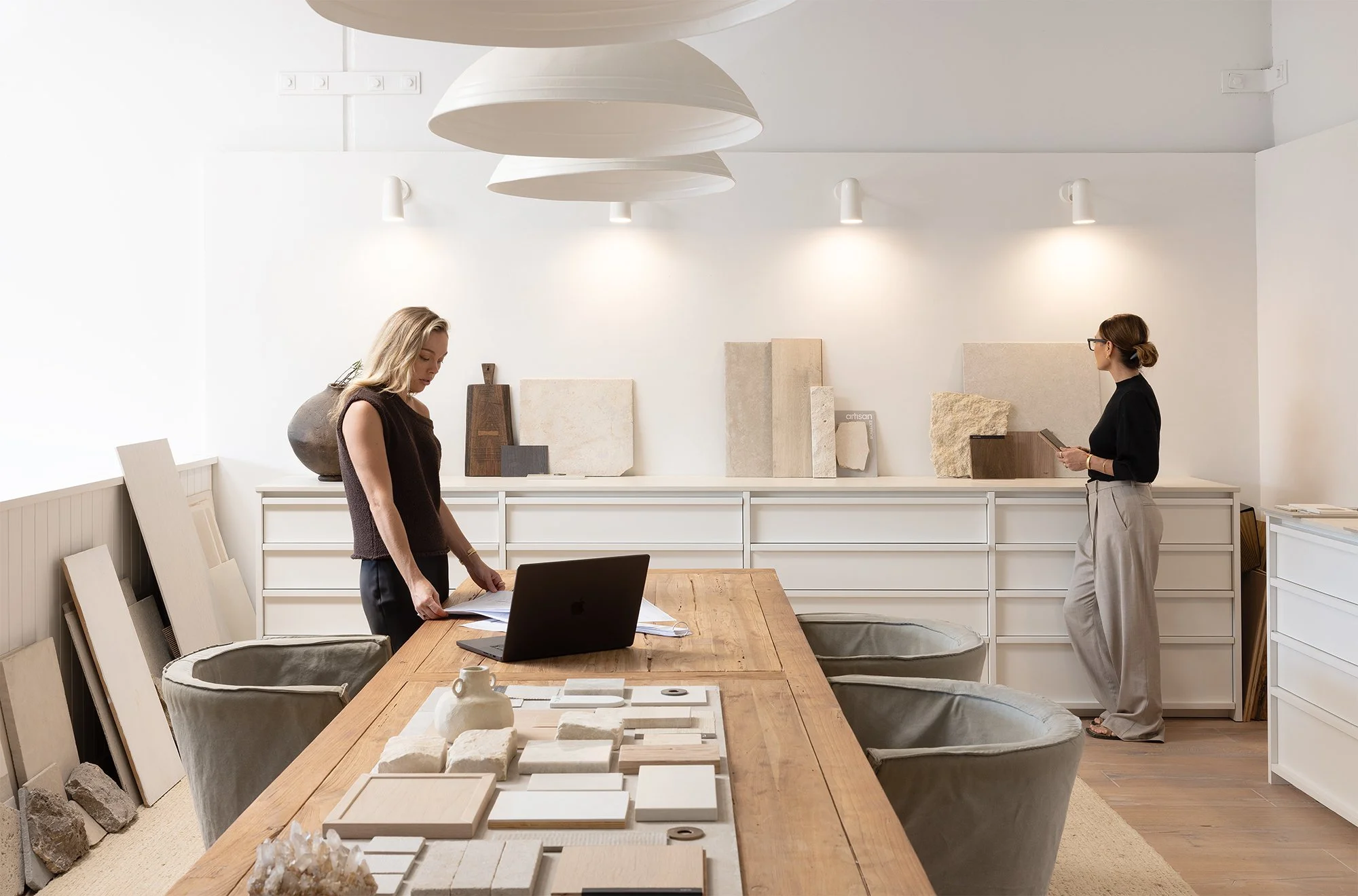 Two women preparing for a presentation in a bright, minimalistic room with samples of tiles and décor on shelves and a wooden table with a laptop and papers.