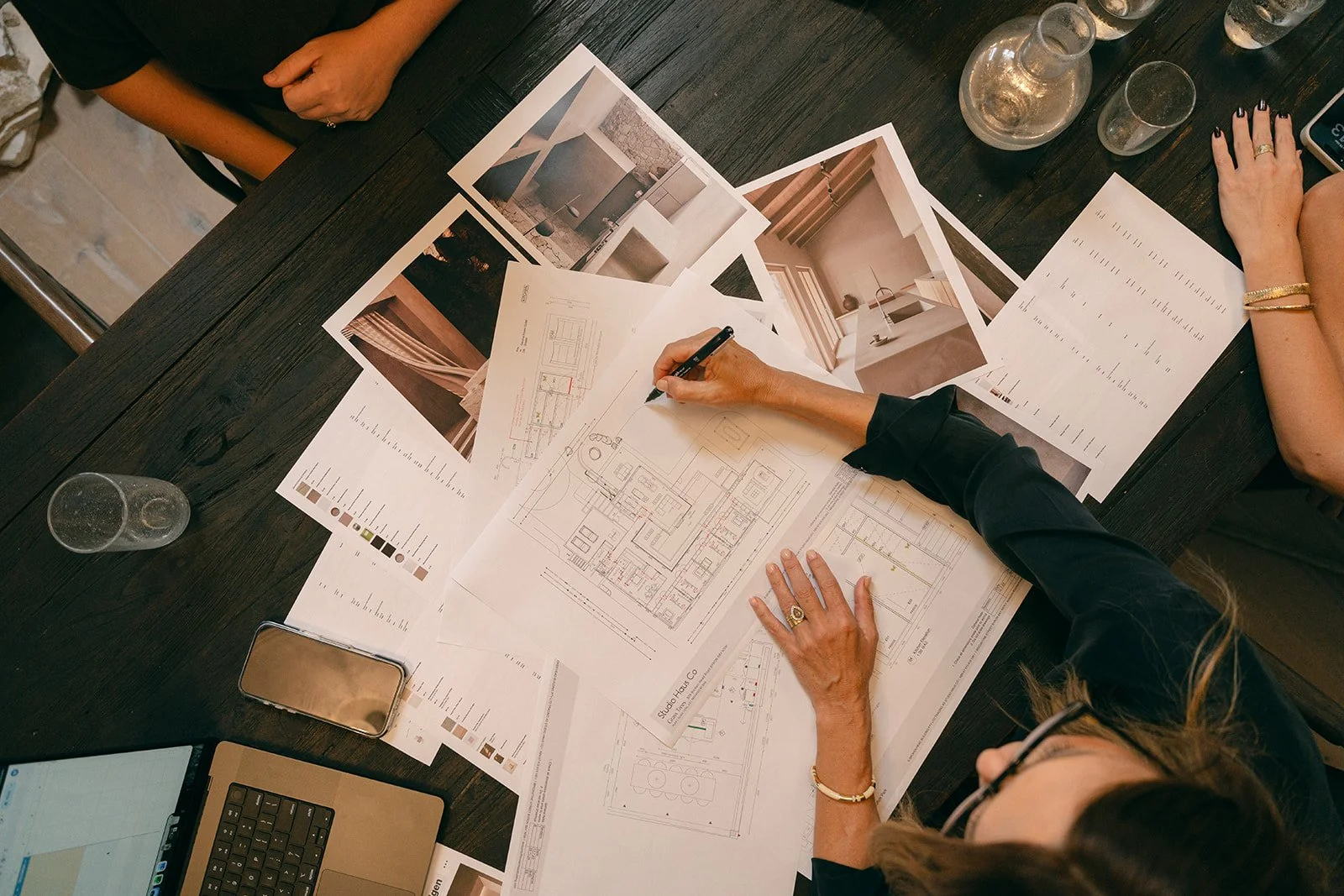 Architectural plans and interior design images spread across a dark wooden table with two people working on drawings, surrounded by water glasses, a smartphone, and a laptop.
