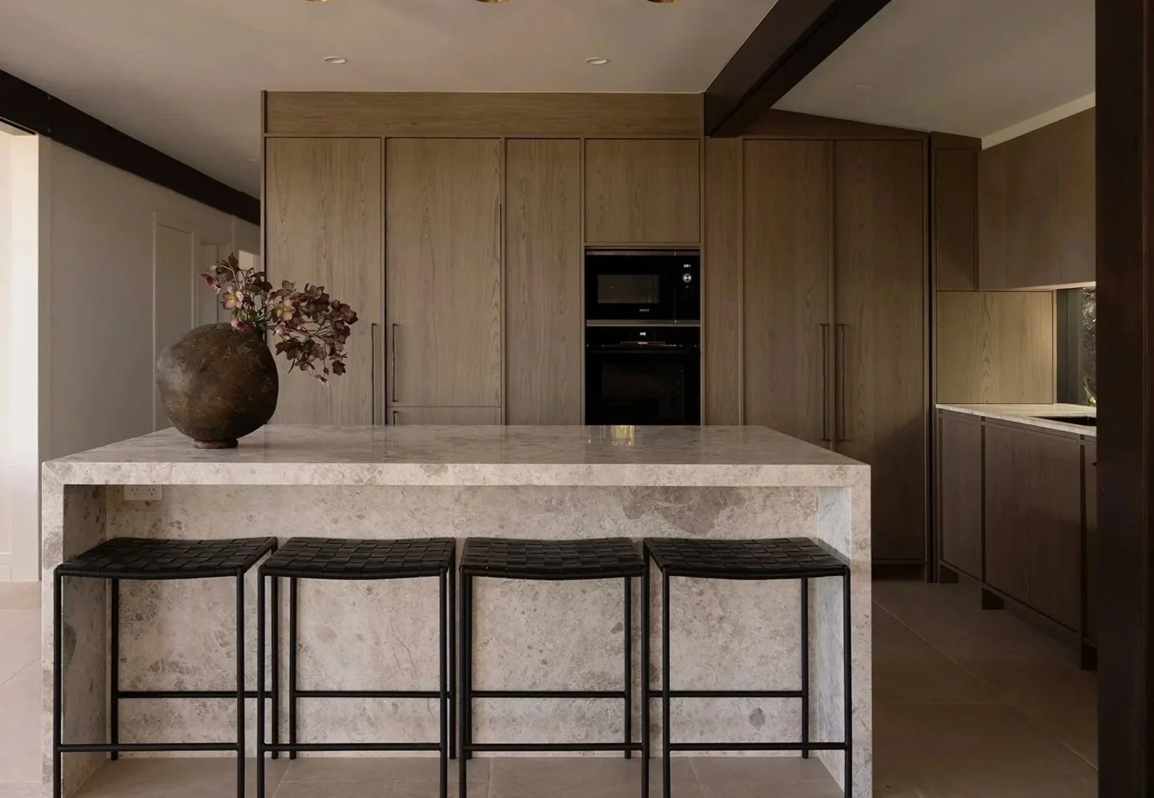 Modern kitchen with a large marble island, three black leather stools, wooden cabinets, a black oven and microwave, a ceramic vase with branches on the island, and a window near the sink.