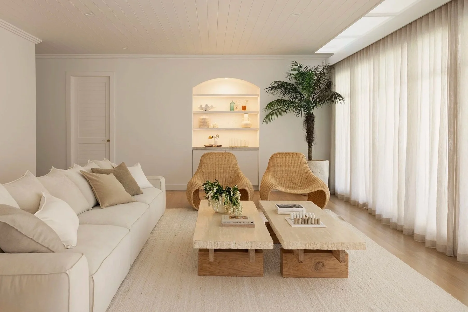 Living room with beige and white color scheme, featuring a white sofa with beige and brown cushions, a light wood coffee table with books and a flower arrangement, two wicker chairs, a large potted palm tree, sliding sheer curtains, and built-in wall shelves with decorative items.