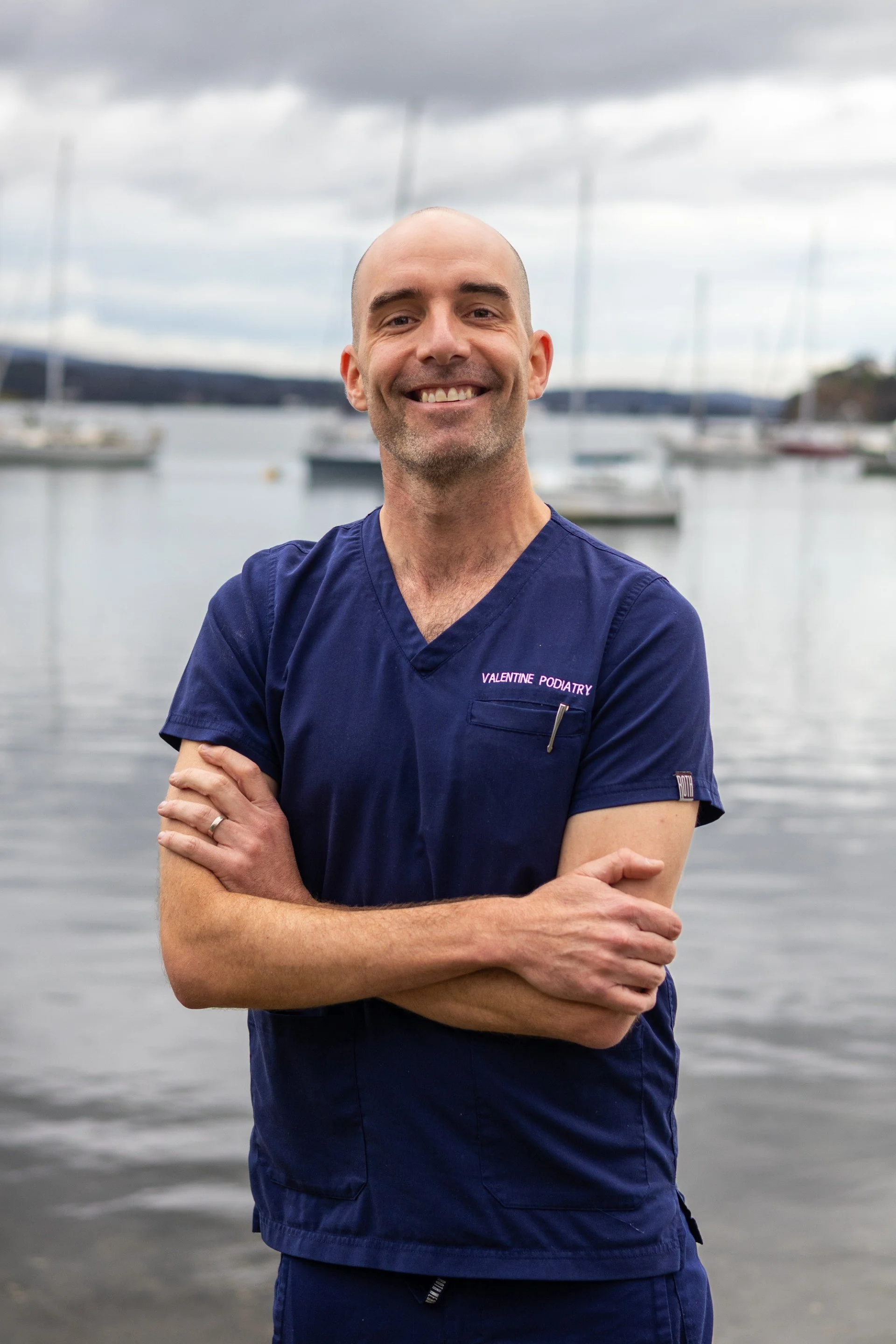 A man with a bald head and light beard smiling, wearing a dark blue medical scrub with 'Valentine Podiatry' embroidered on it, standing indoors with a plain white wall and ceiling tiles in the background.