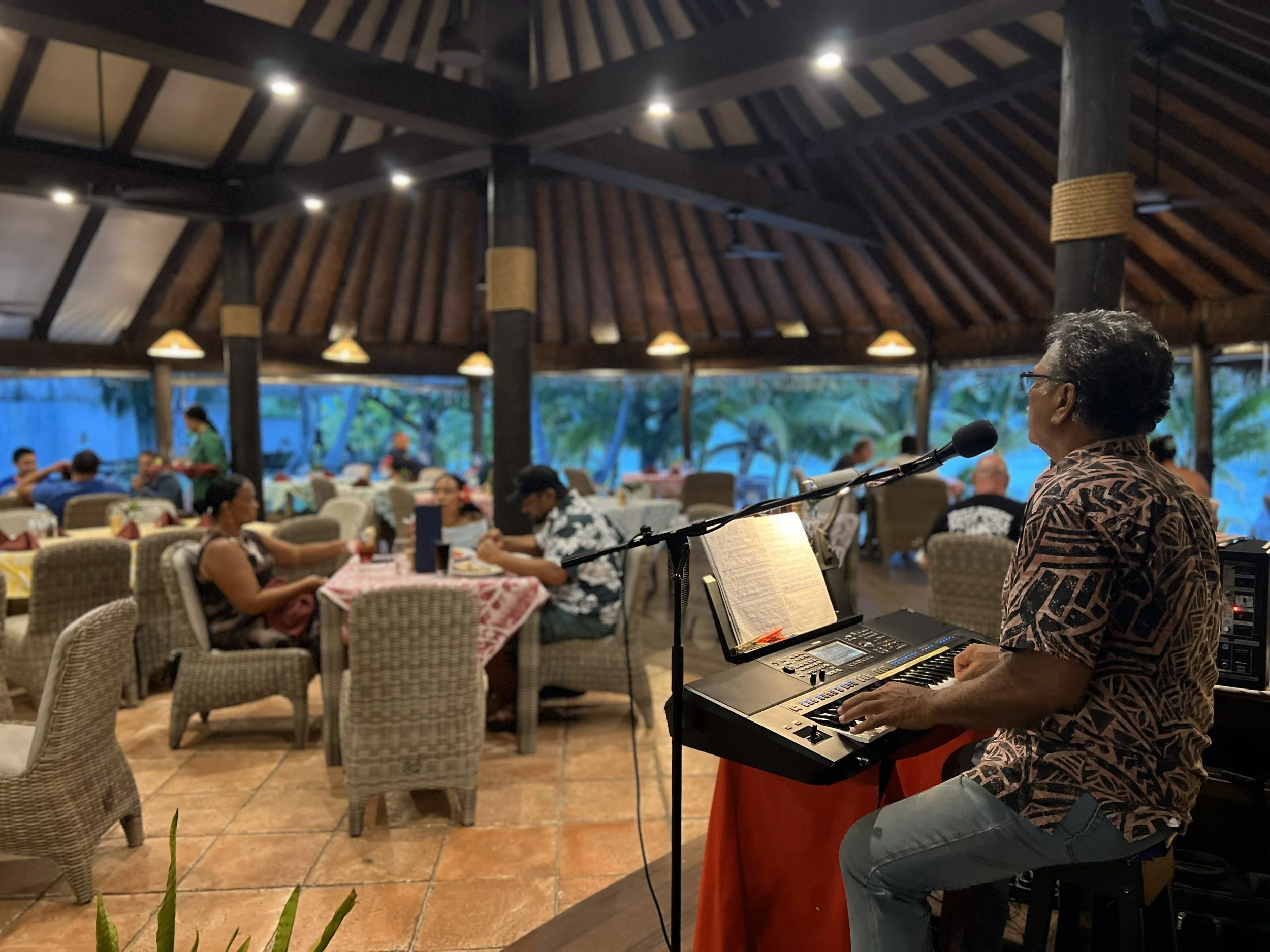 A musician playing keyboard and singing into a microphone in a restaurant with diners seated at tables, with large windows showing palm trees outside.