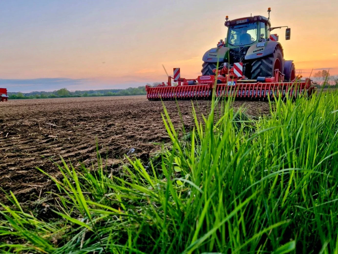 Ein Traktor mitAnhängern auf einem Acker bei Sonnenuntergang, mit grünem Gras im Vordergrund.