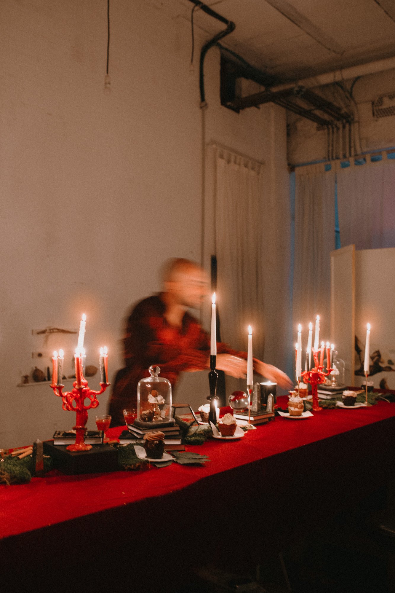 Mesa decorada con candelabros y cupcakes, entre cortinas y tuberías en un ambiente interior, con una persona moviéndose rápidamente.