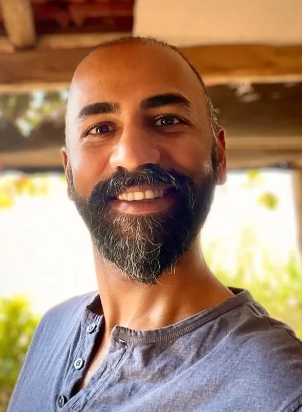 A smiling man with a beard and mustache, wearing a gray shirt, standing outdoors with trees and a wooden structure in the background.