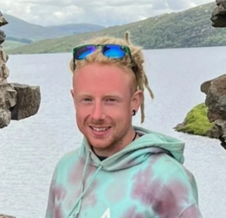 A man with blonde dreadlocks, sunglasses on his head, and a hoodie standing near a body of water with rocky landscape and green hills in the background.