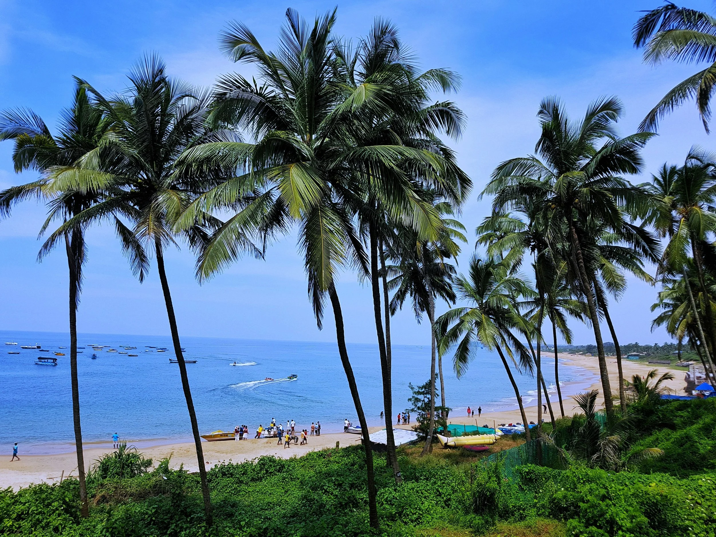 Beach scene with palm trees in the foreground, people walking on the sandy shore, boats in the water, and a clear blue sky.