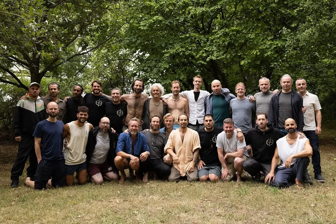 A group of 24 men gathered outdoors on grass, with trees in the background, posing for a photo.