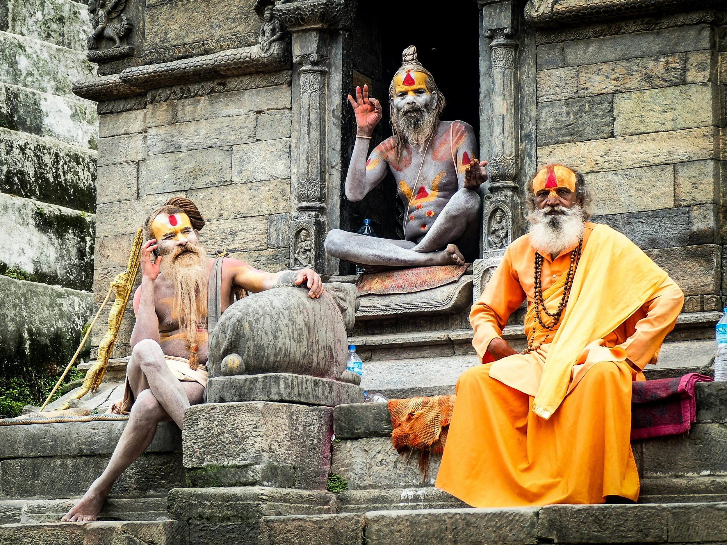 Three Hindu Sadhus sitting and posing for the camera on stone steps outside a temple. They are painted with traditional markings on their foreheads and bodies, and are dressed in saffron-colored robes. The main Sadhu in the center is sitting in a cross-legged position, making a gesture with his right hand and holding a small object in his left. The other two are seated nearby, one with a phone to his ear. The temple is made of stone with intricate carvings.