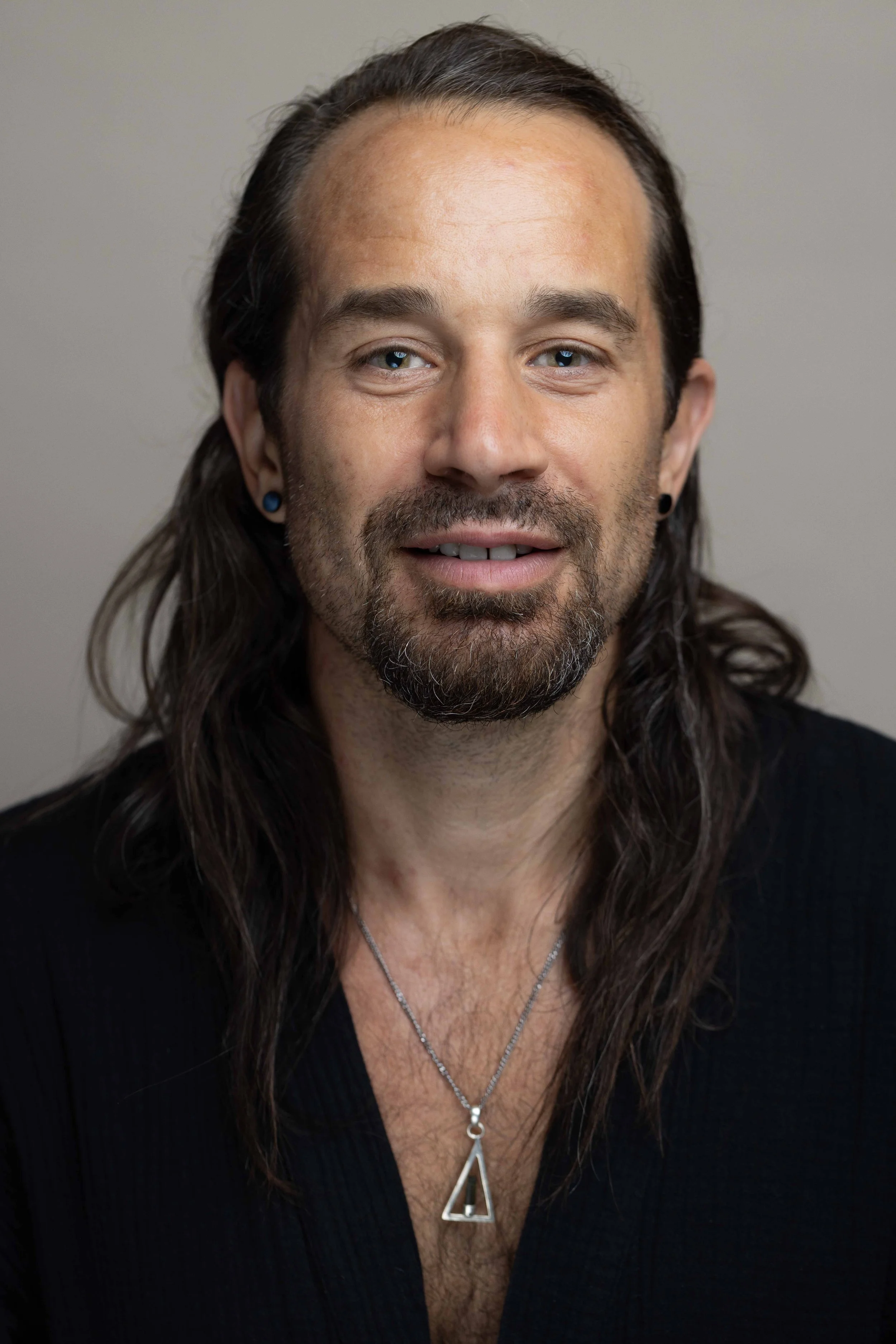 Close-up portrait of a man with long brown hair and blue eyes, wearing a black top, two necklaces, and earrings, looking at the camera against a plain background.