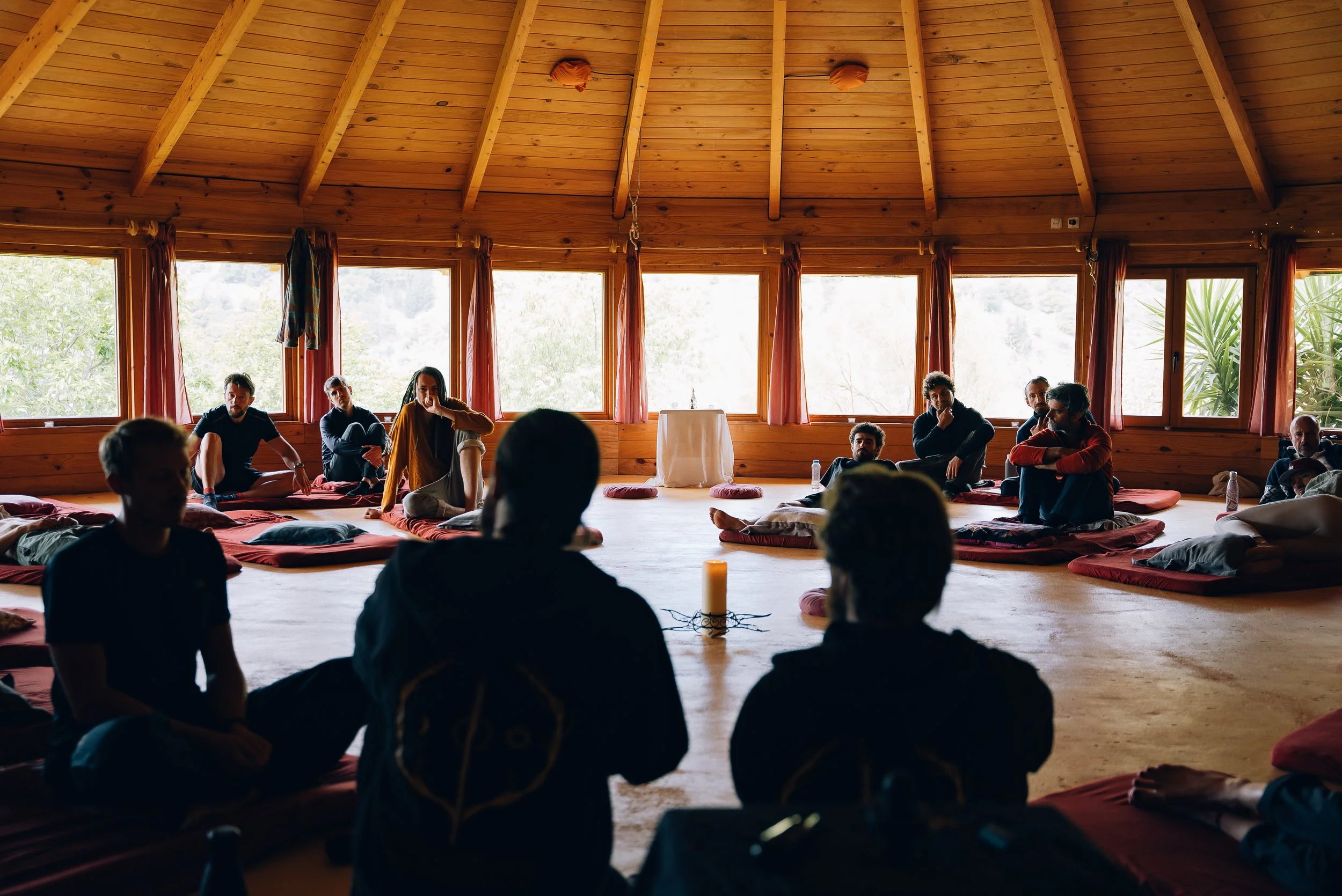 People sitting on cushions in a wooden room with large windows, during a group meditation or workshop.