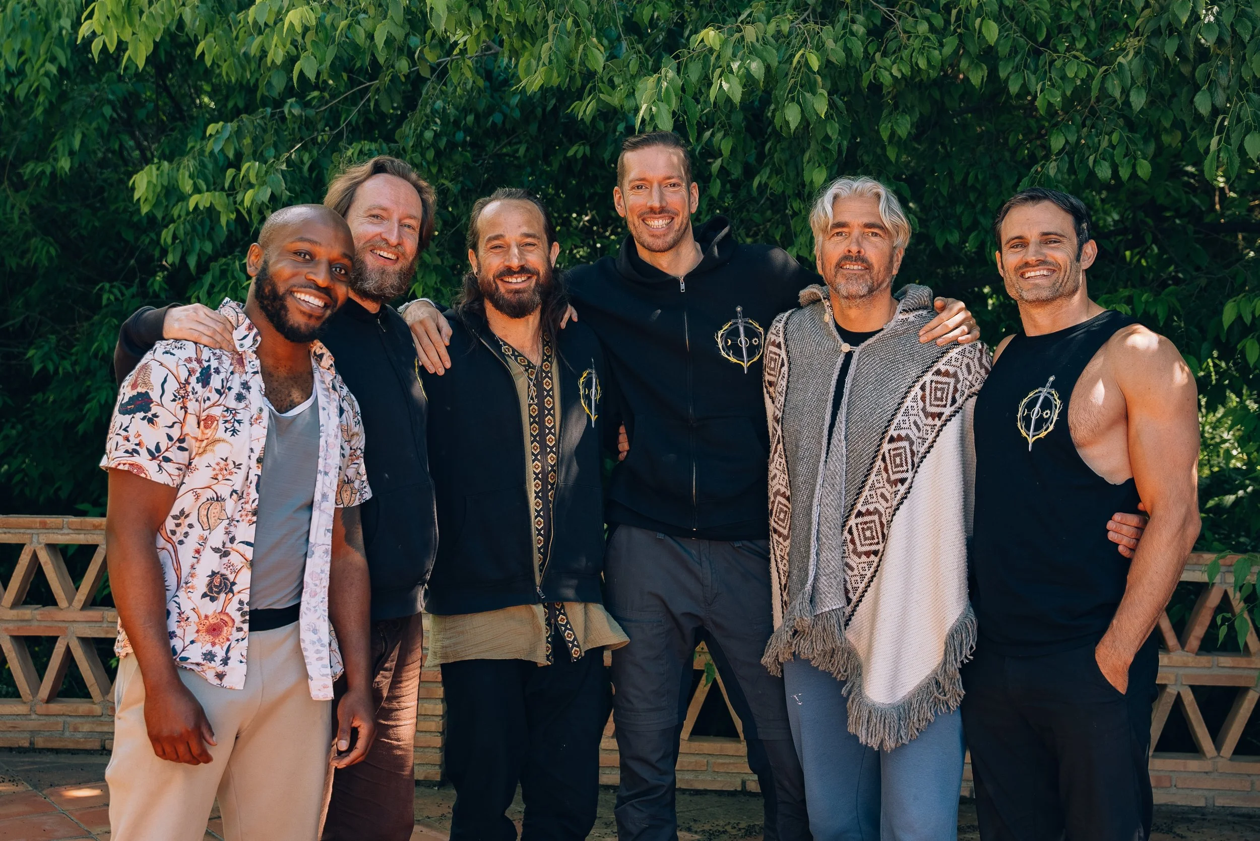 Seven diverse men standing together outdoors in front of leafy green trees, smiling and posing for the photo.