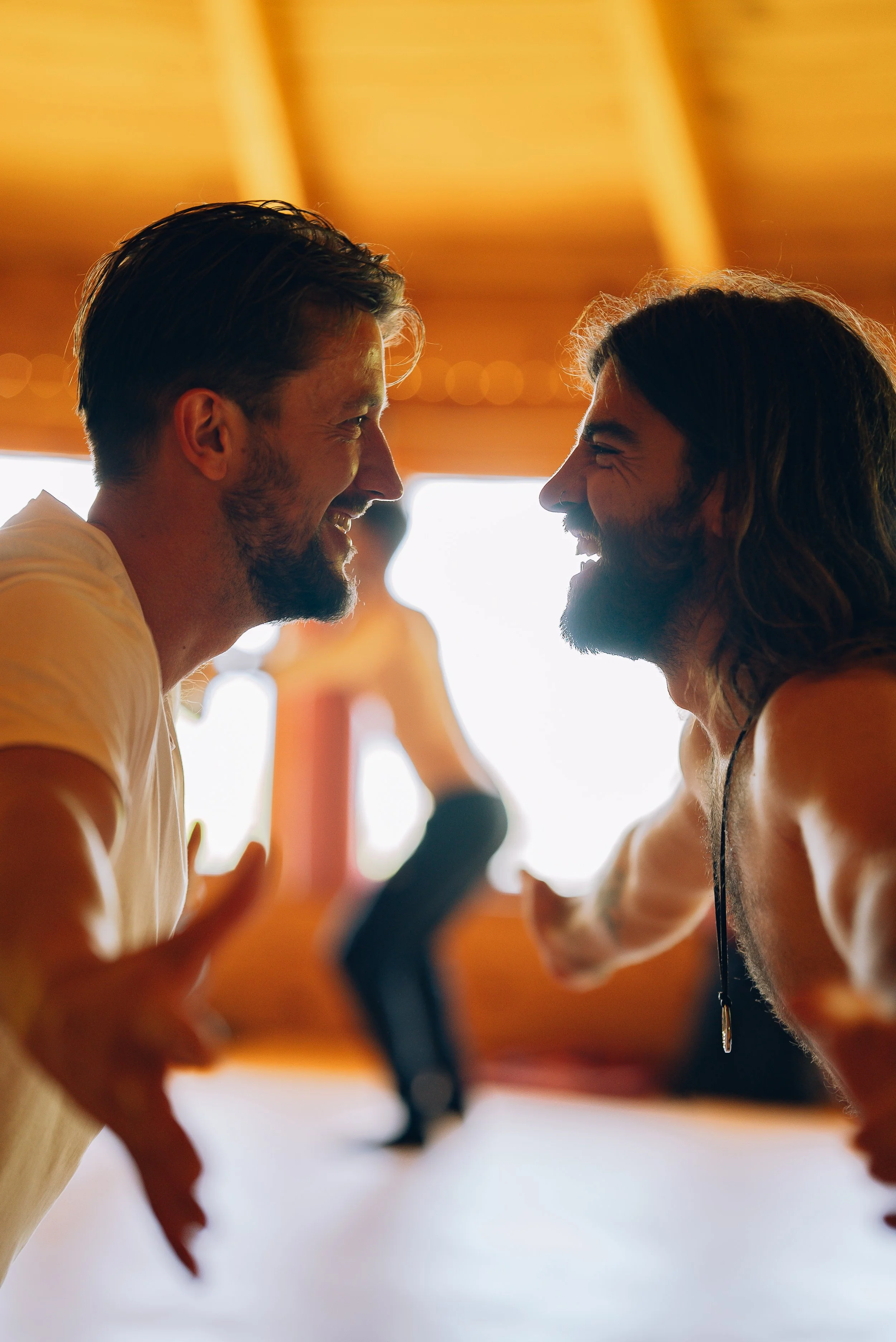 Two men with long hair and beards smiling and facing each other, with a warm yellow background.