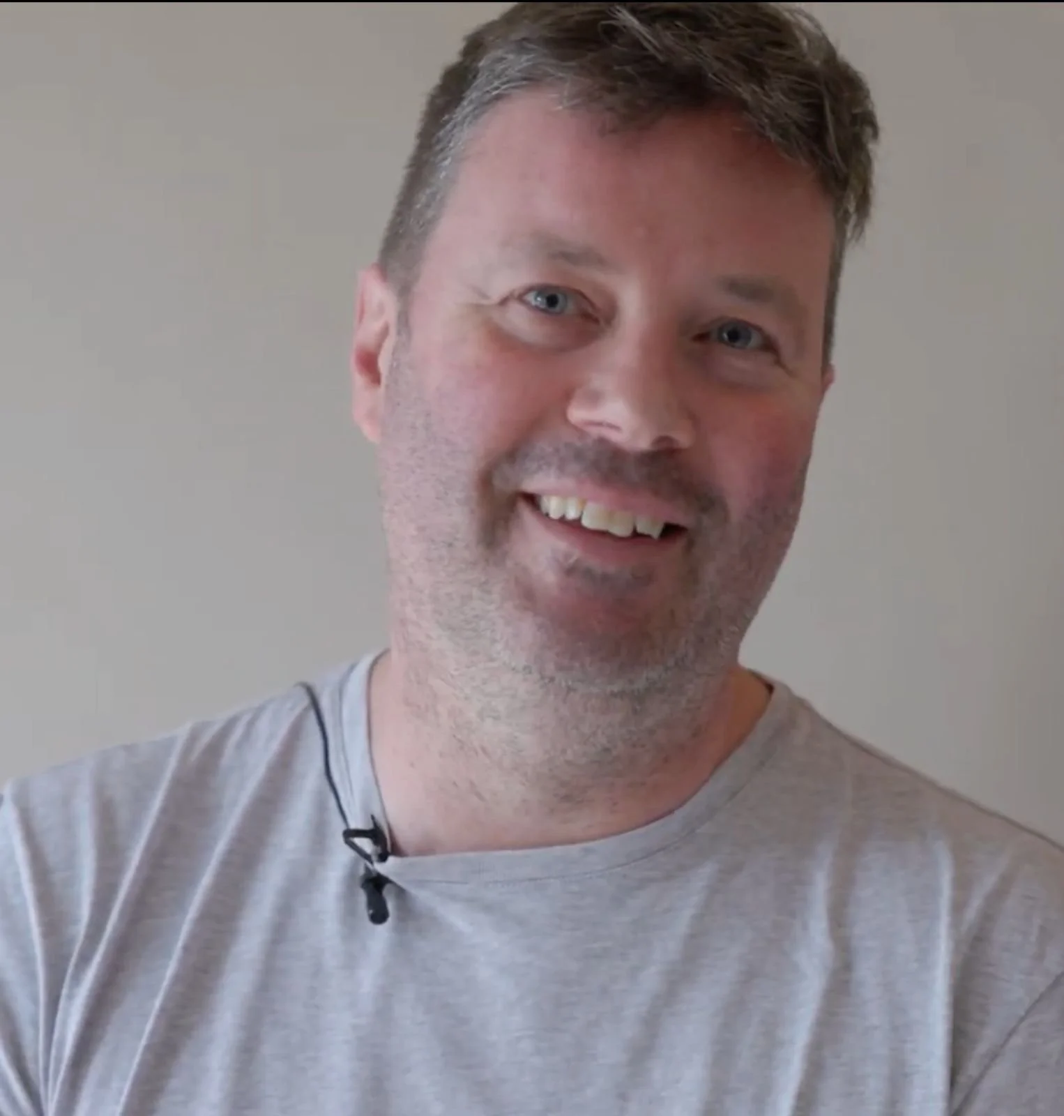 A man with short, brown hair and blue eyes, smiling and wearing a light gray T-shirt with a small black microphone clipped to the collar, standing against a plain light-colored wall.
