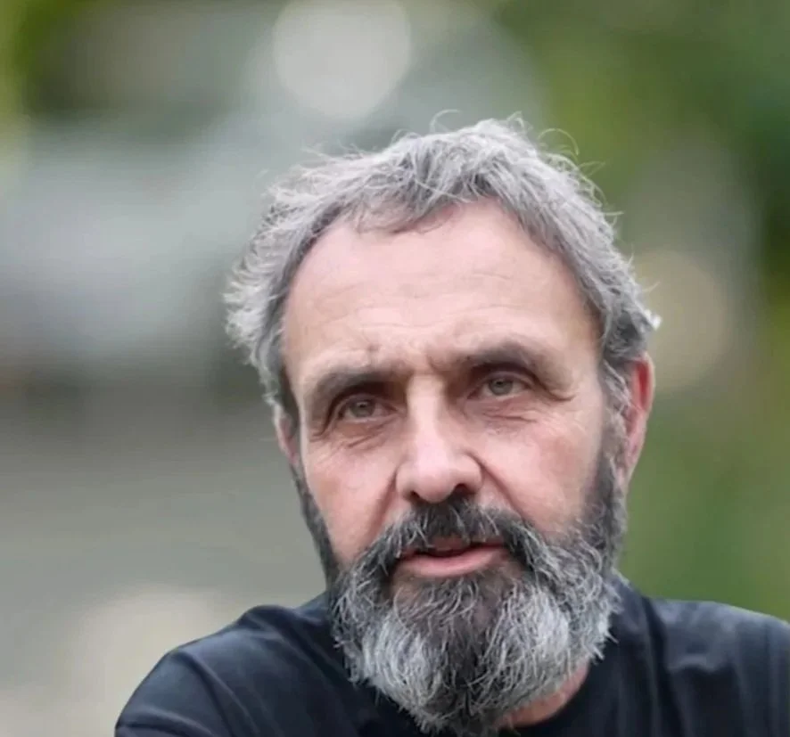 A middle-aged man with gray hair and a beard looks to the side outdoors with a blurred green background.