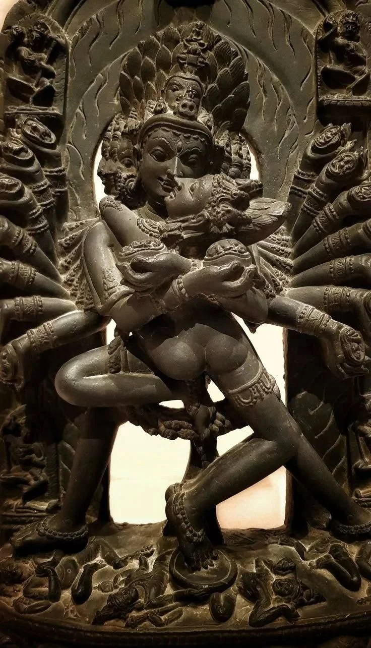 A detailed black carved sculpture of a multi-armed deity holding a smaller figure, with multiple heads and intricate adornments, surrounded by additional smaller figures.