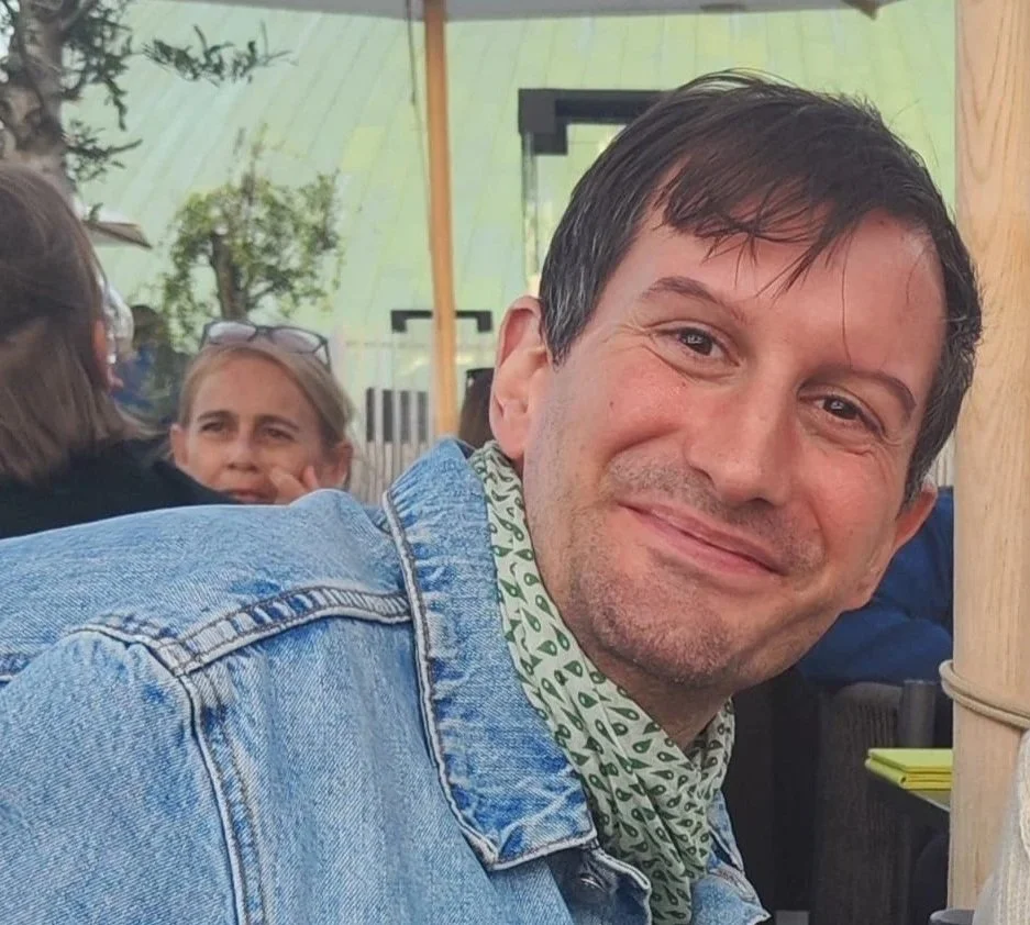 A man with dark hair and a light beard, smiling, wearing a denim jacket and a patterned scarf, sitting at an outdoor event with other people in the background.