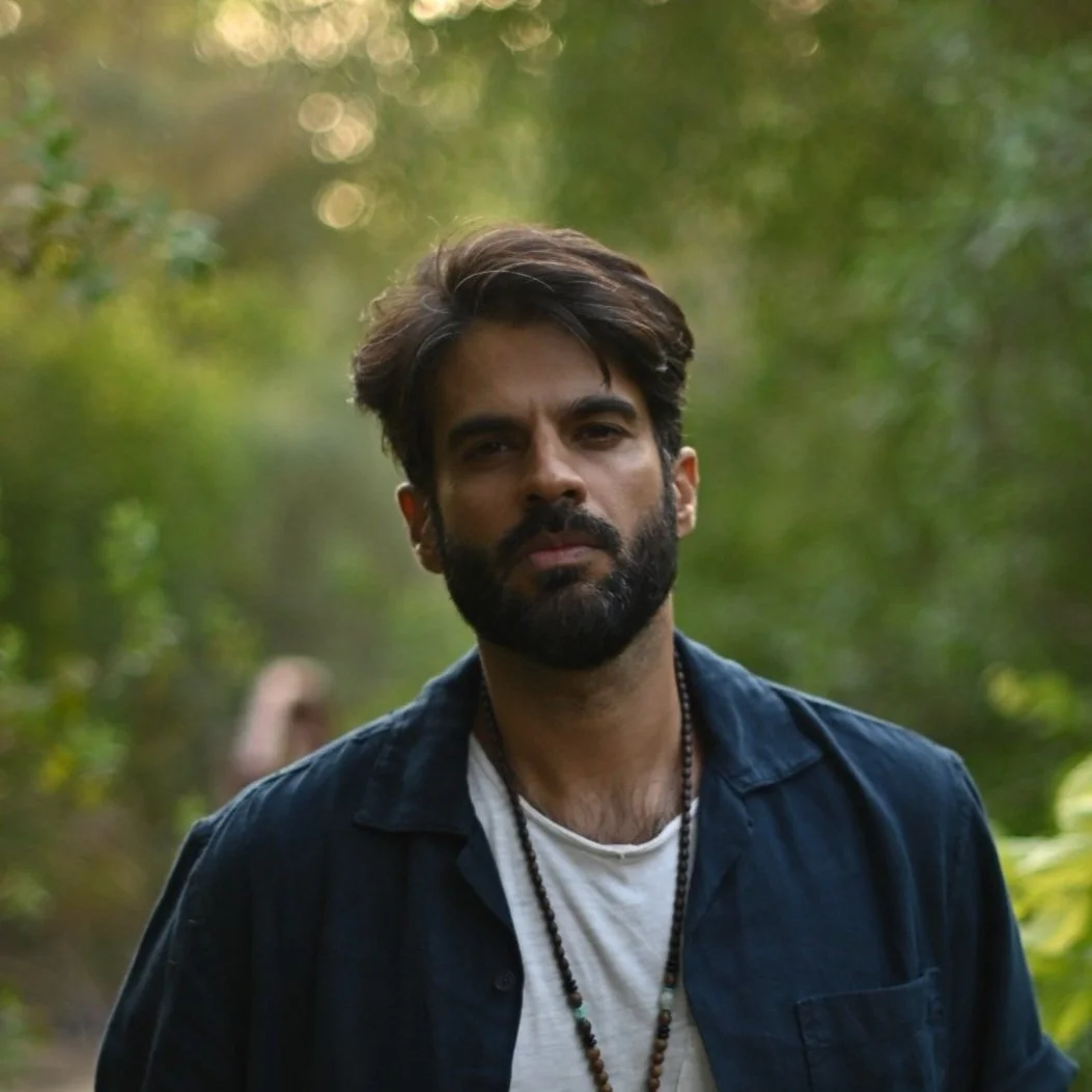 A man with a beard and styled hair stands outdoors in a green, blurred natural environment. He is wearing a dark jacket over a white shirt, with a beaded necklace. He looks directly at the camera with a neutral expression.