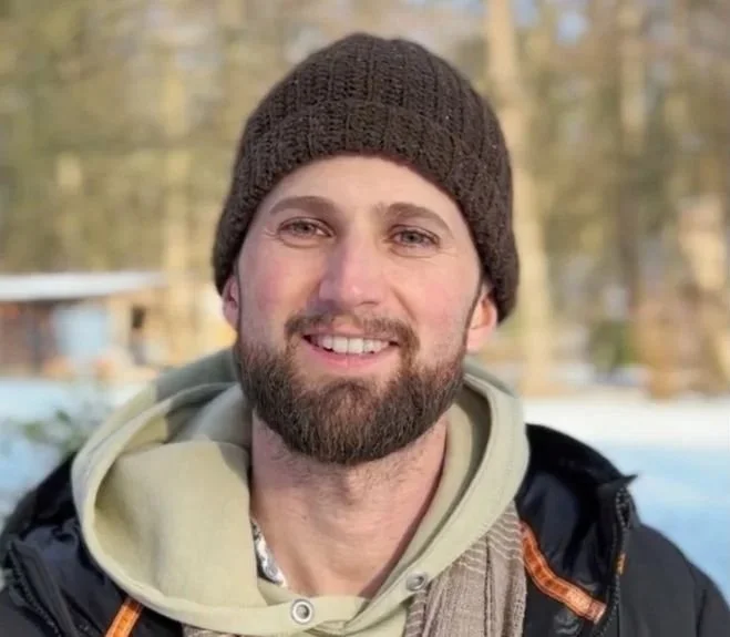A man outdoors wearing a brown knit beanie, beige hoodie, and a black jacket with an orange accent, smiling at the camera, with a snowy background and trees.