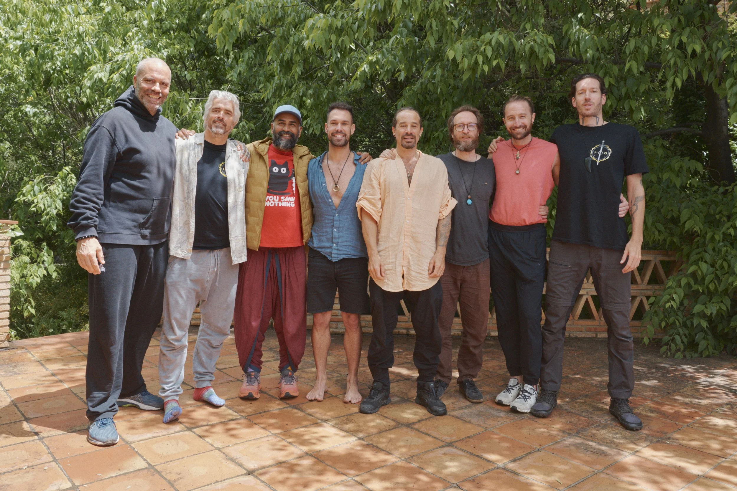 A group of nine men standing together outdoors on a brick patio, surrounded by green trees. The men are smiling and have their arms around each other's shoulders, dressed casually in t-shirts, hoodies, and shorts.